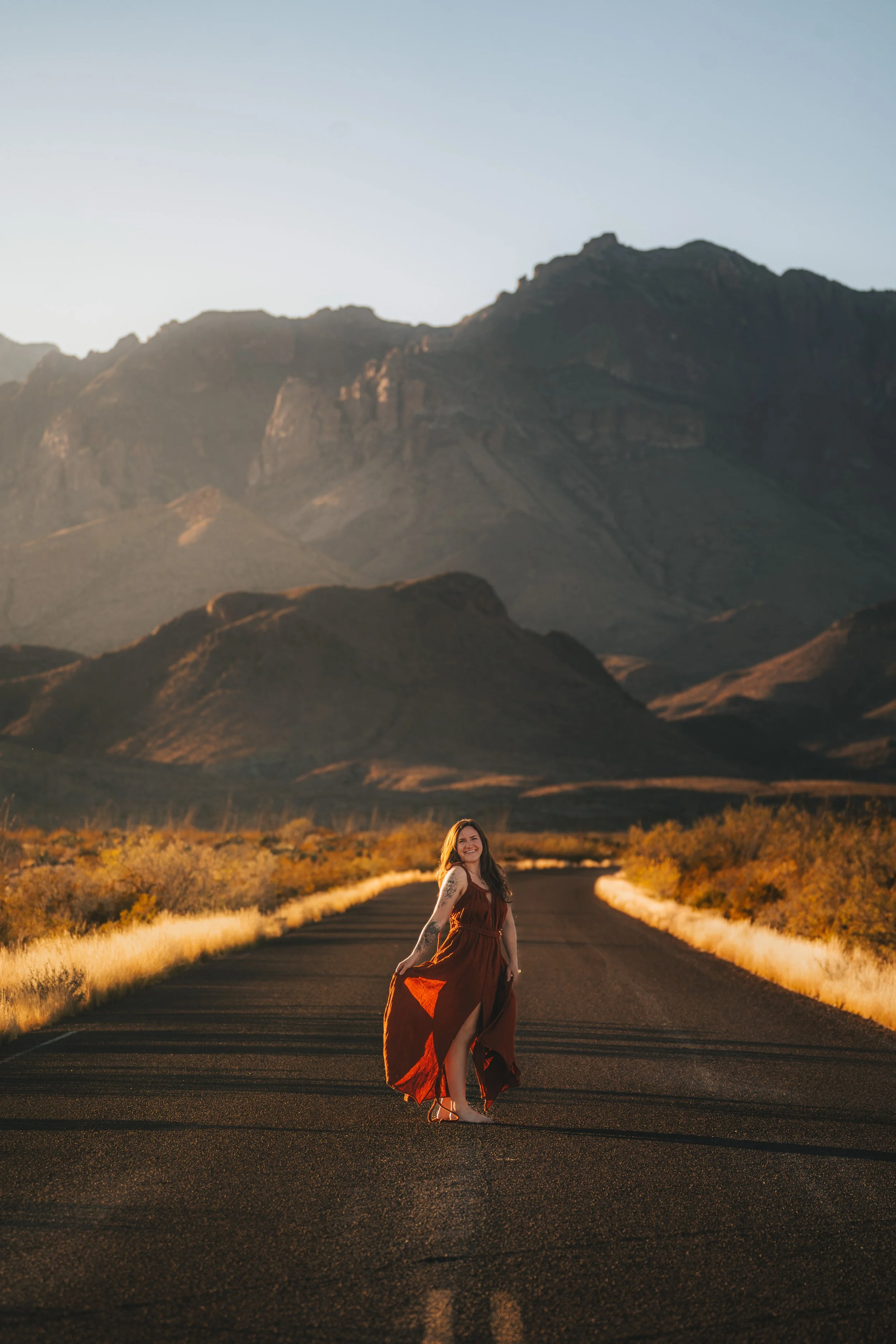 A woman in a brown dress standing on a deserted road with a mountainous landscape in the background during golden hour.