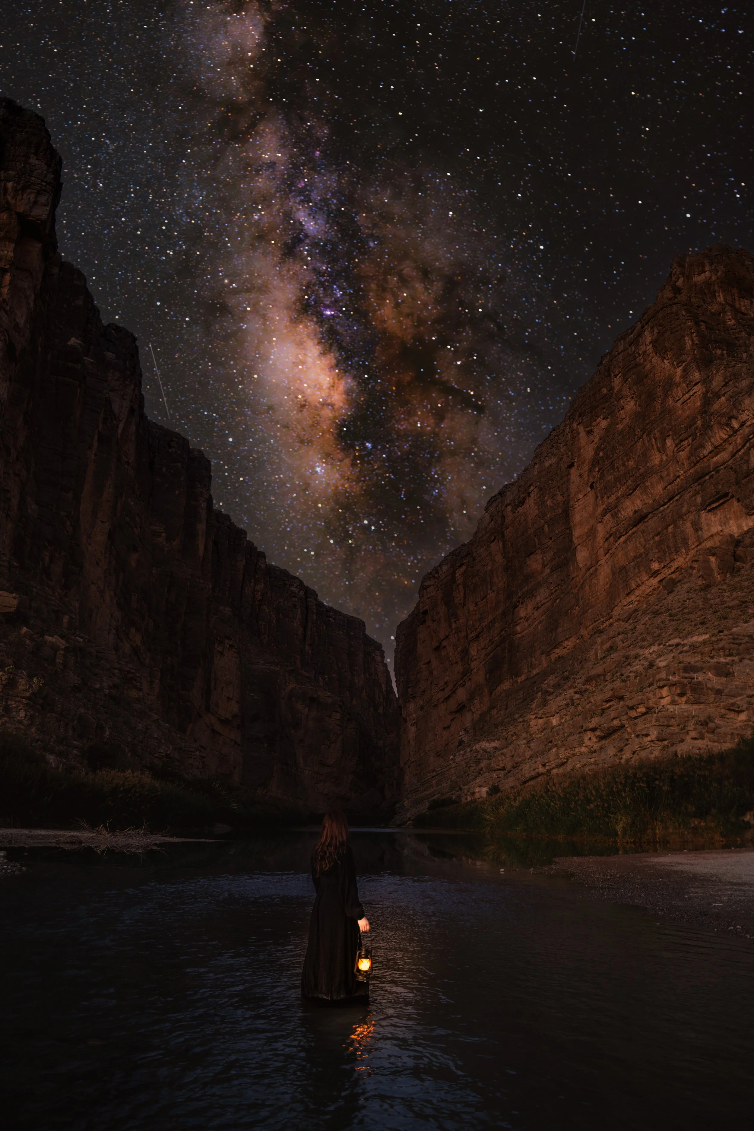A woman standing in a river at night, holding a lantern, with towering canyon walls on either side and a starry sky with the Milky Way overhead.