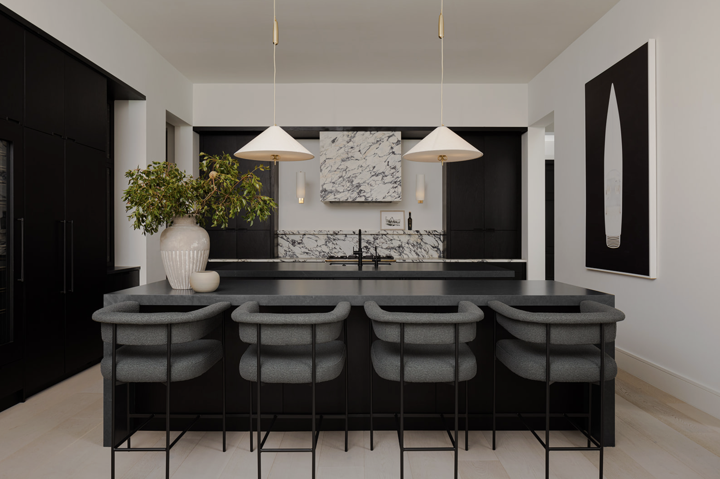 Modern kitchen with black cabinetry, marble backsplash, and gray barstools at a black island with a large potted plant and minimalist art. Taffe McArdle Design