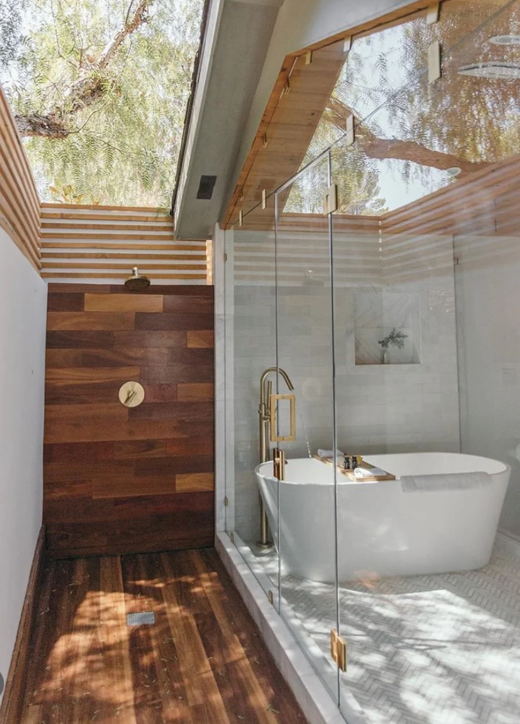 Modern bathroom with a freestanding bathtub, glass shower enclosure, and wooden accents. Large window allows natural light and shows tree branches outside.