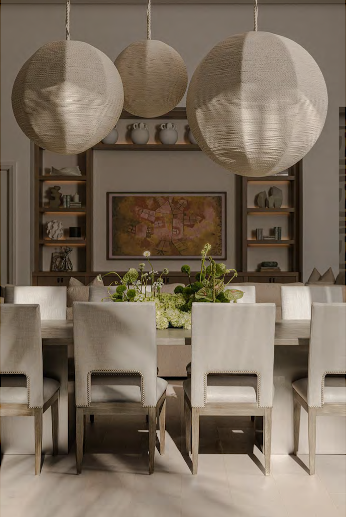 A dining room scene featuring a long table with cream-colored chairs, a floral centerpiece, three large hanging paper lanterns, a built-in bookshelf with decorative items, and a colorful framed painting on the wall.