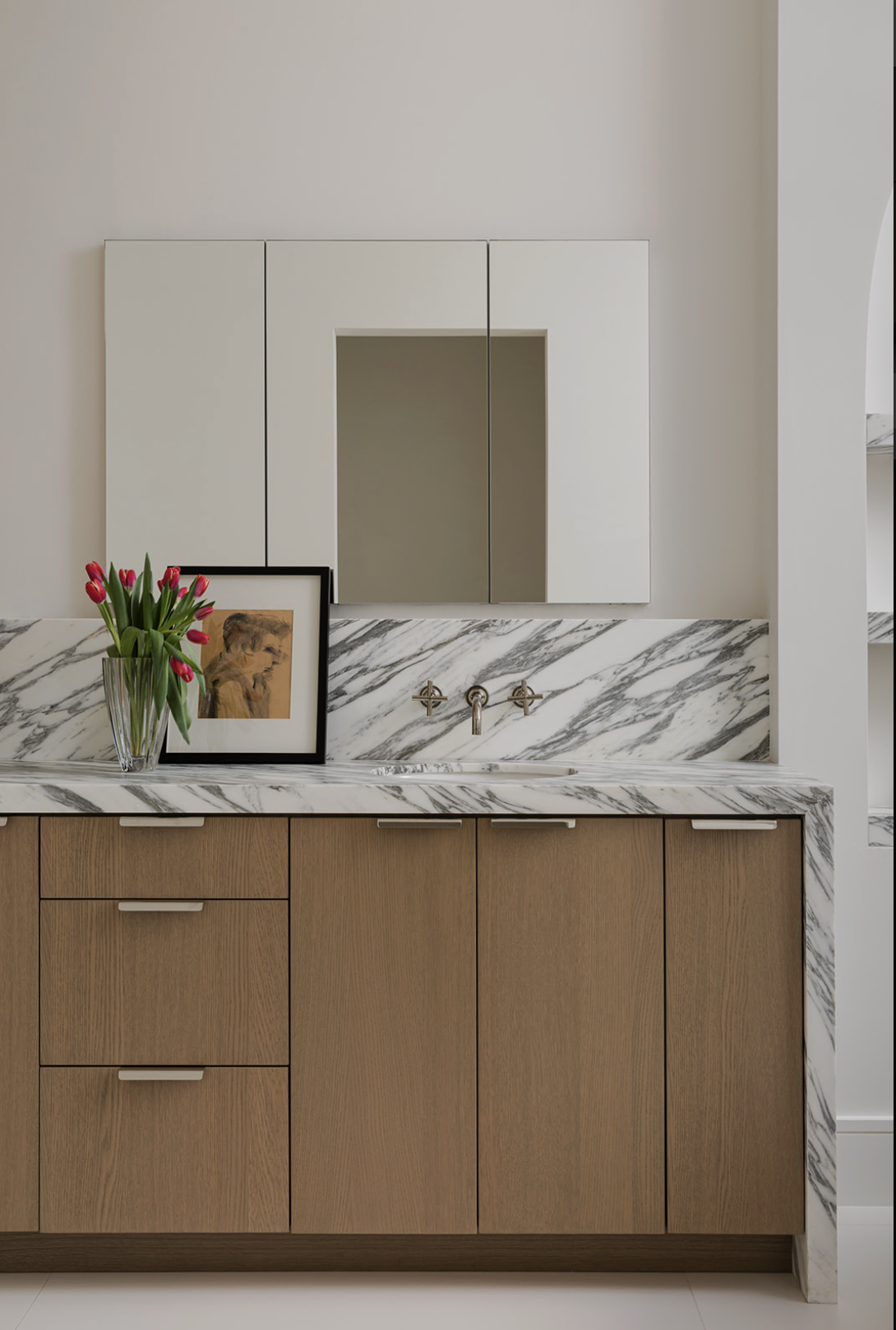 A bathroom vanity with a marble countertop, wooden cabinets, and a mirror cabinet above. A vase of pink tulips and a framed sketch are on the counter.