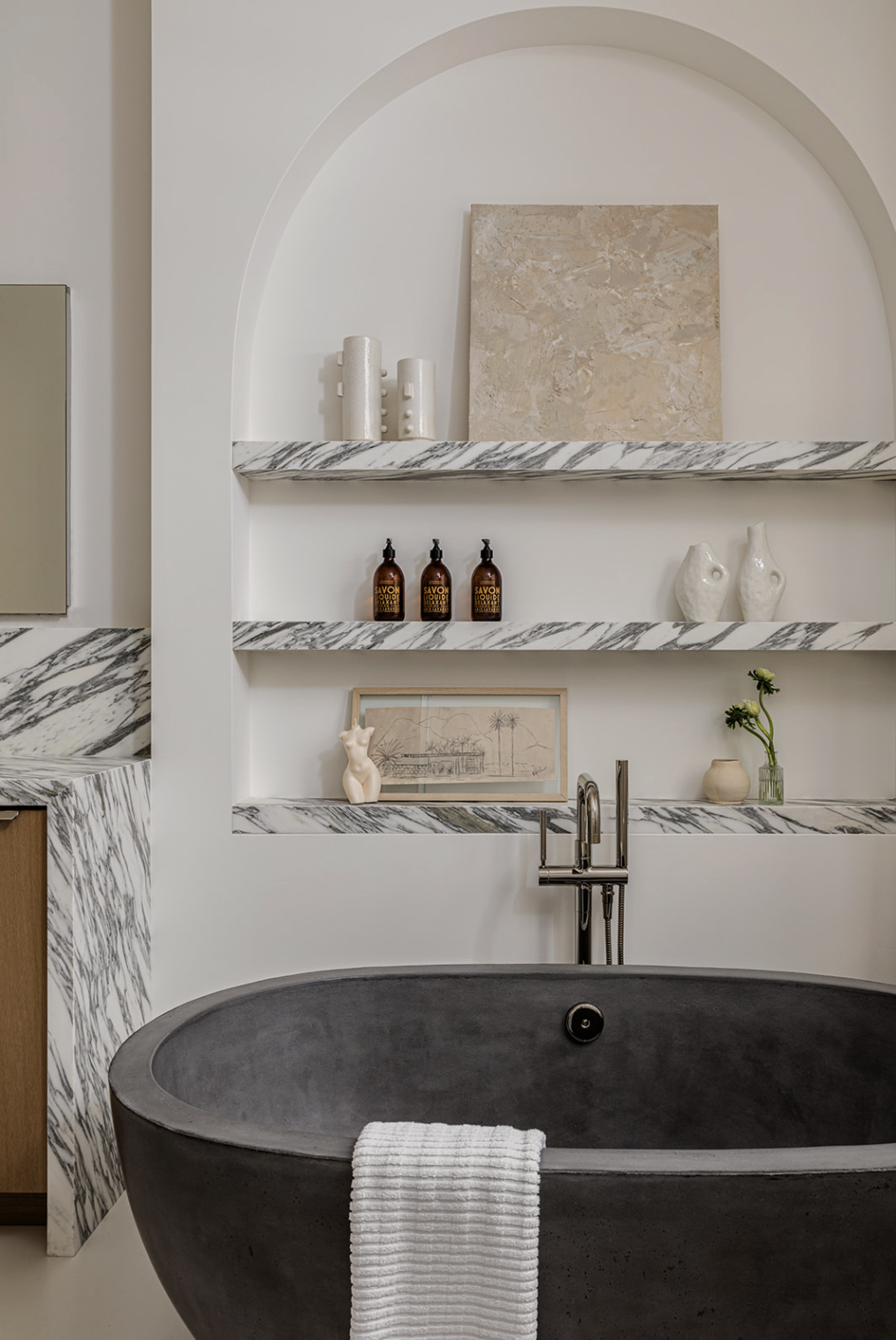 Modern bathroom with a black bathtub, marble shelves, and decorative items including vases, framed art, and candles.
