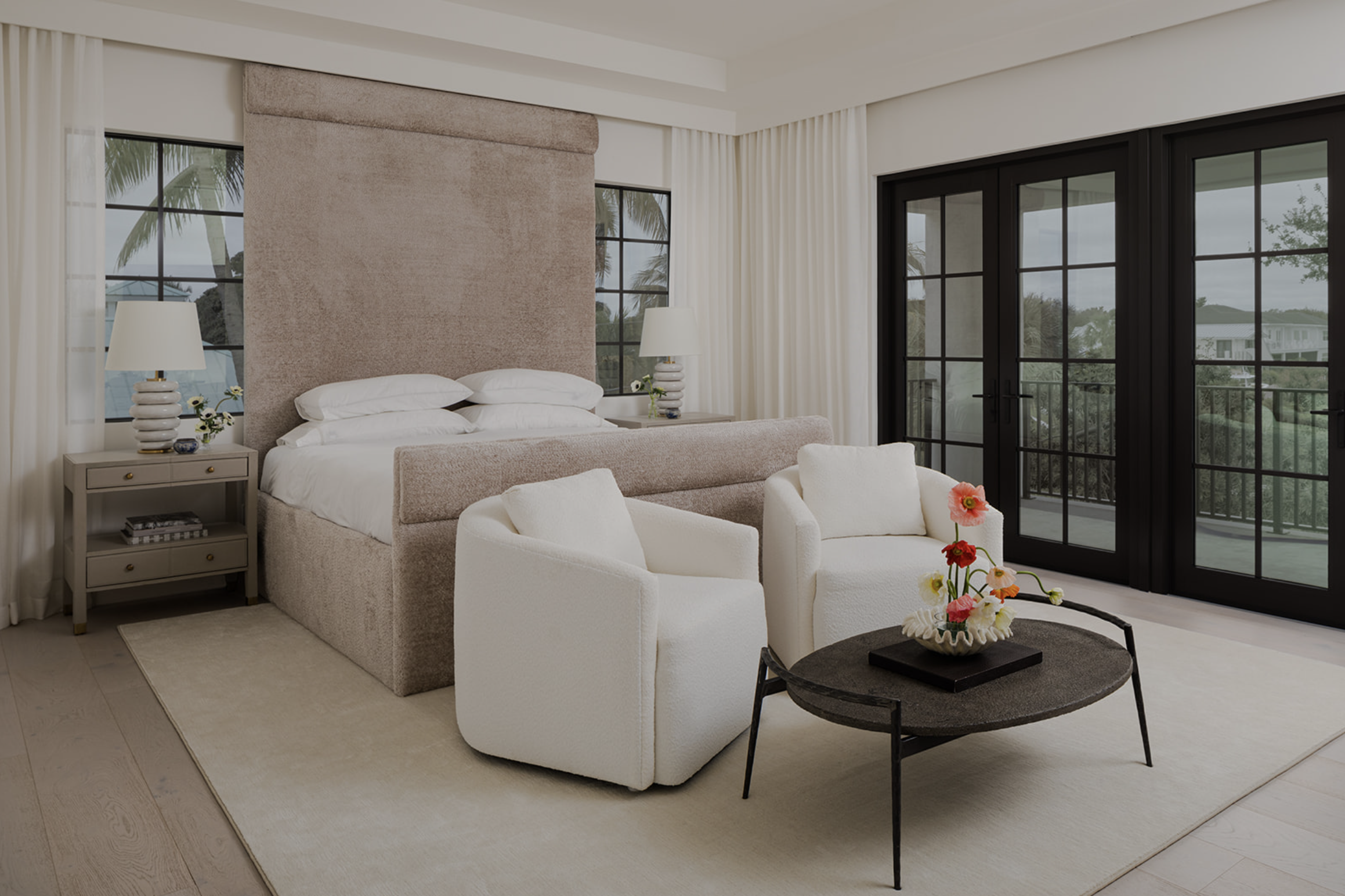 A luxurious bedroom with a large bed, white pillows, two bedside tables with lamps, white armchairs, a round coffee table with a flower arrangement, and large glass doors leading to a balcony with a view of trees and houses.
