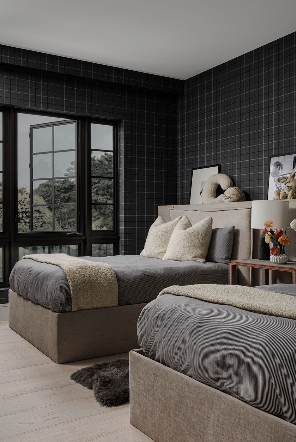 A modern bedroom with twin beds, dark window frames, black grid-patterned walls, beige upholstered headboards, gray bedding, cream-colored pillows, a small bedside table with a flower vase, and decorative items on the headboard.