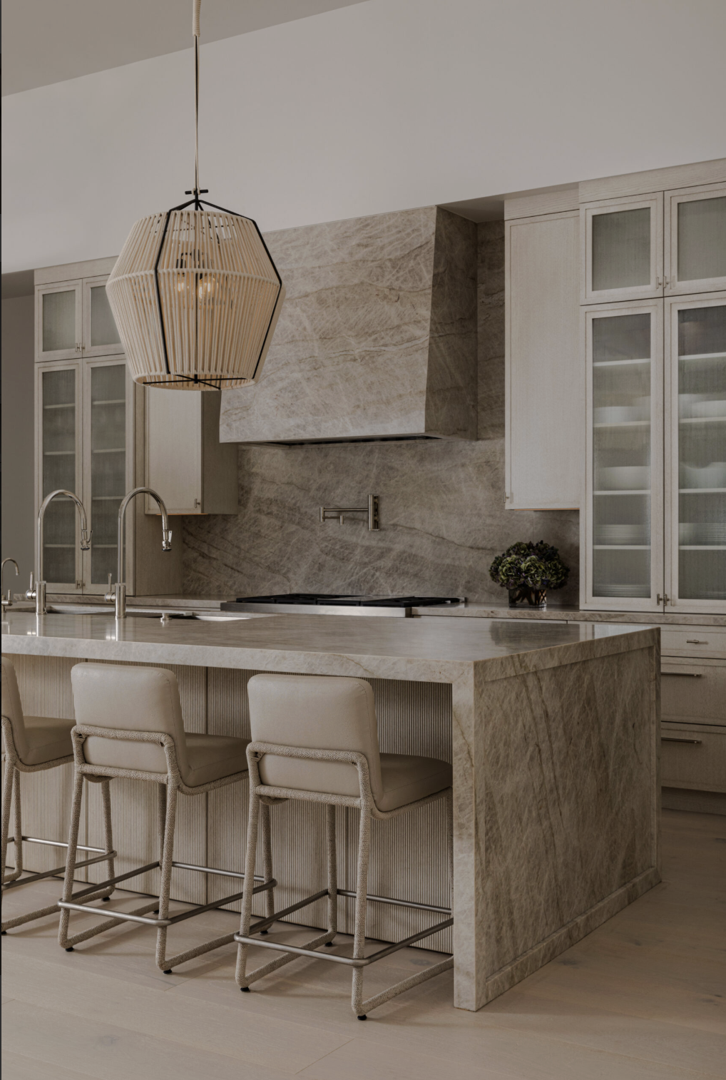 Modern kitchen with marble island, bar stools, glass-front cabinets, and a geometric wooden pendant light.