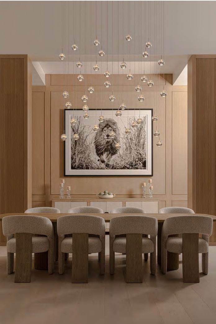 A dining room with a large wooden table and eight beige upholstered chairs. The room has wooden wall panels, a black and white lion photograph, and a modern chandelier with hanging glass globes. Taffe McArdle Design