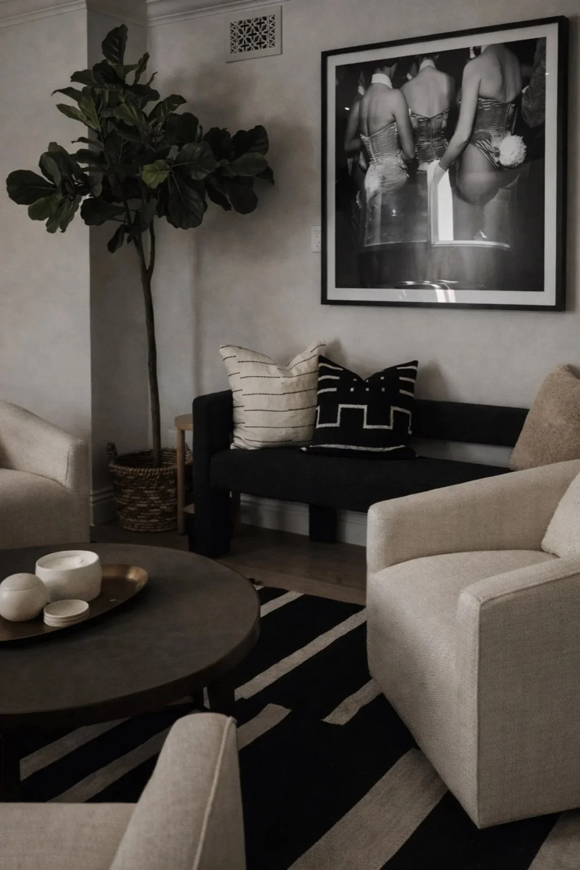 Living room with beige armchair, black sofa with black and white pillows, round wooden coffee table with ceramics, large potted plant, black and white photograph on wall.