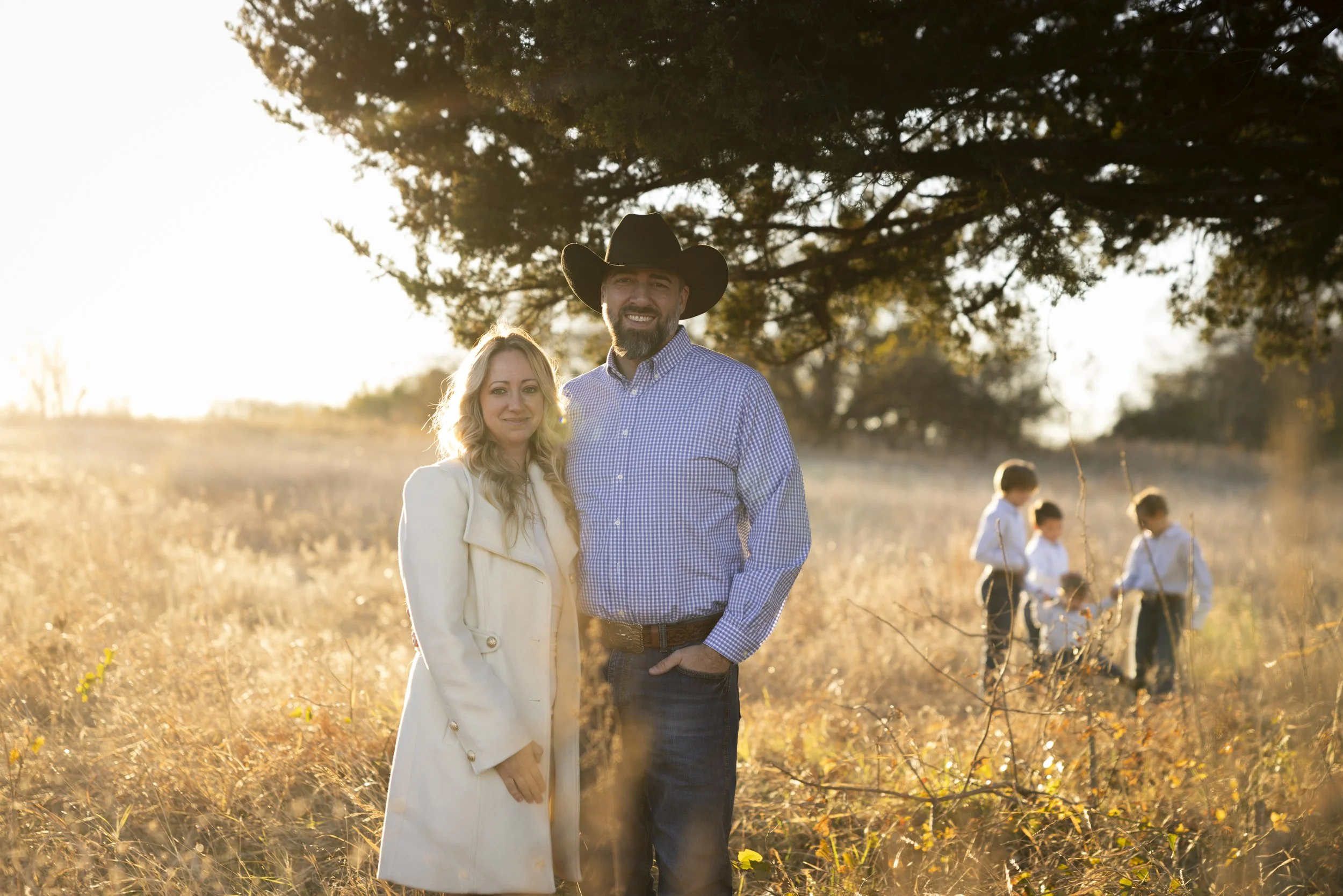 A woman and man standing in a field during sunset, with three children playing in the background under a large tree.