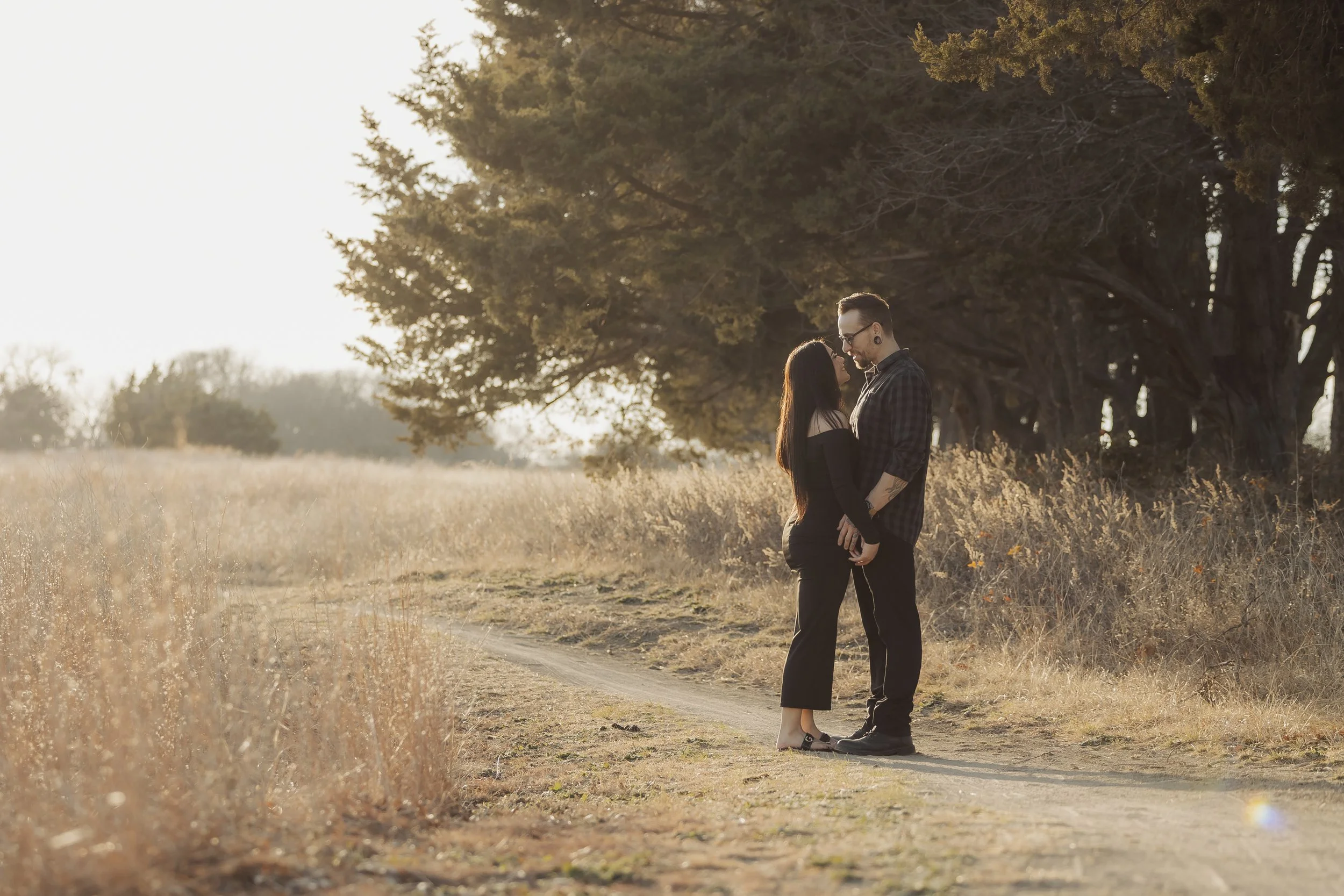 A couple stands close together in a field with tall, dry grass, holding hands and gazing into each other's eyes under a large tree with sprawling branches during sunset.