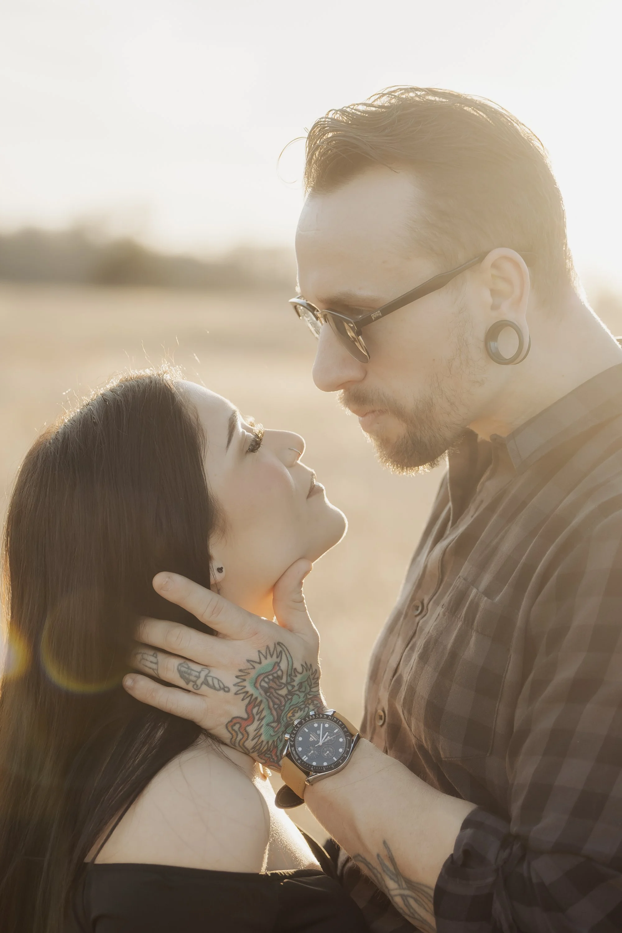 A man and woman in an intimate pose outdoors during sunset, with the man holding the woman's chin and they gaze into each other's eyes.