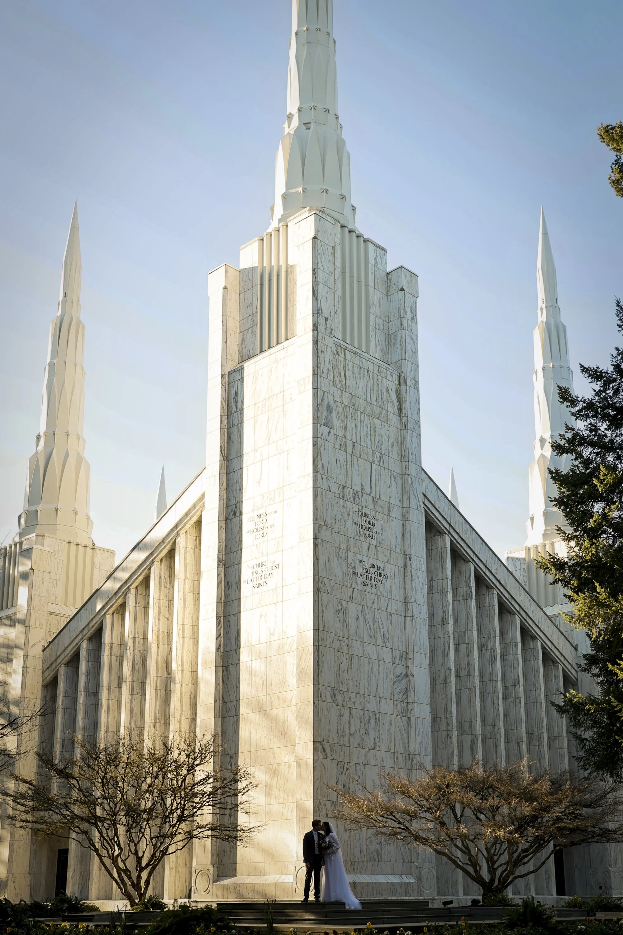 A bride and groom standing close together in front of a large white church with tall steeples under a clear blue sky, with trees on either side.