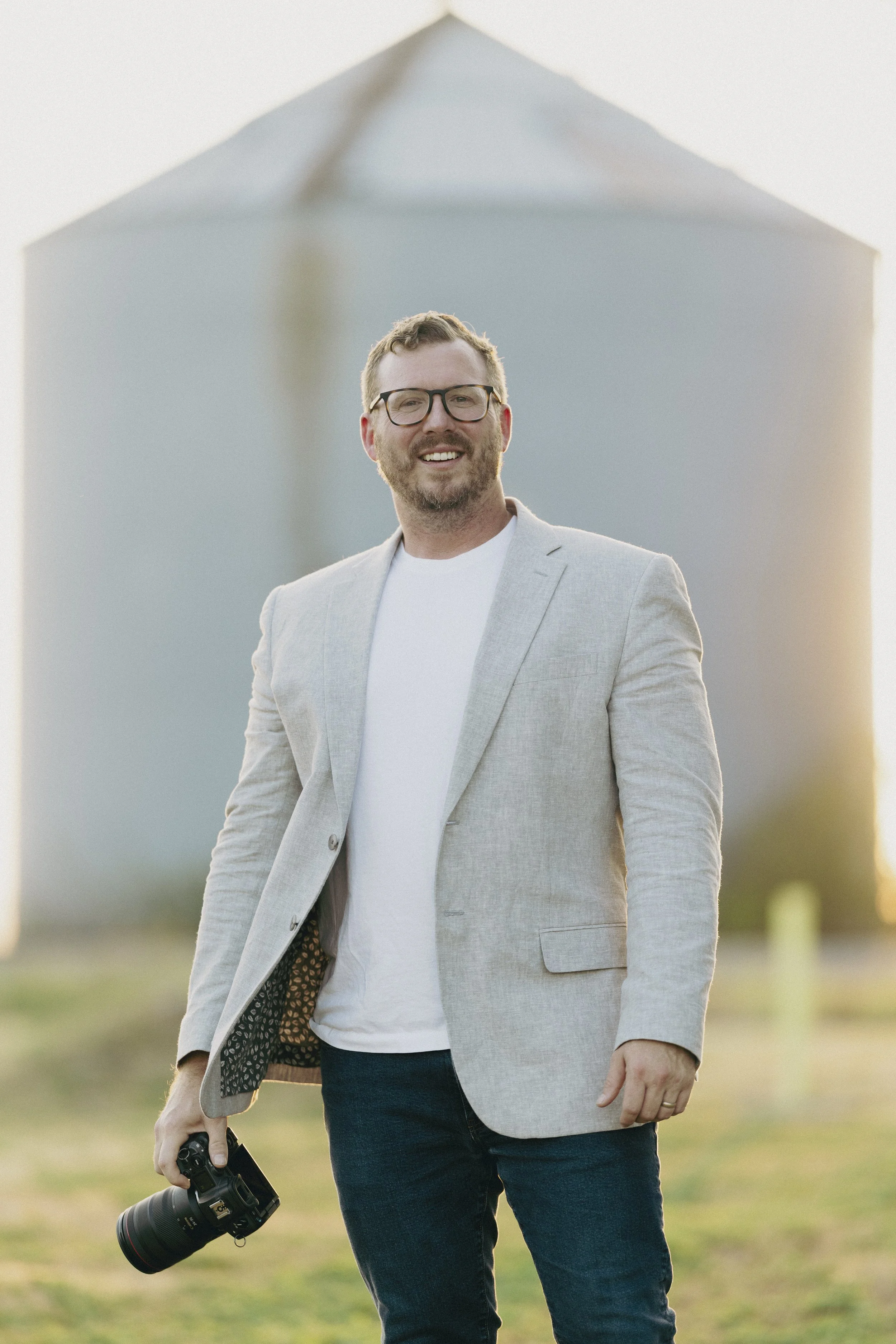 A man with glasses, short hair, and a beard, wearing a light gray blazer and white T-shirt, holding a camera with a large lens in his right hand, standing outdoors with a blurred building in the background.