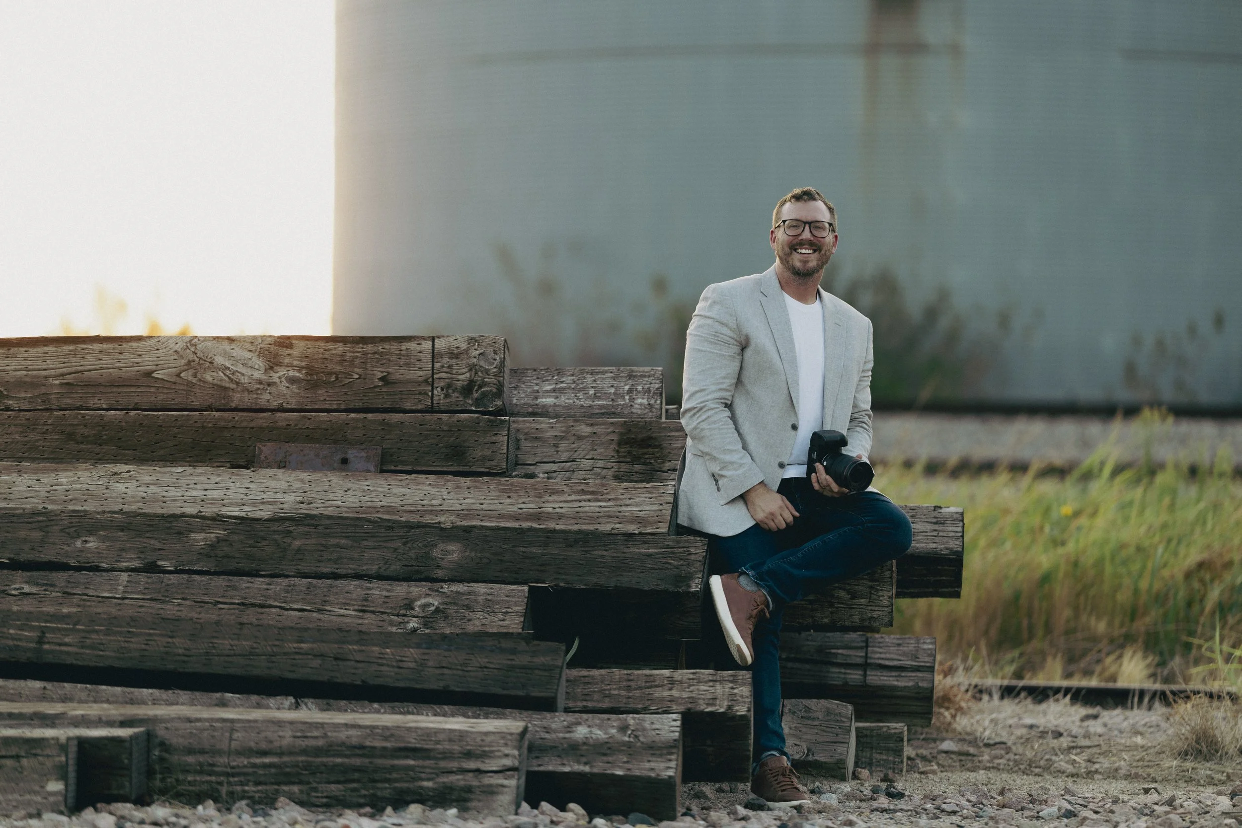 A man with glasses, beard, and short hair, wearing a light gray blazer, white shirt, dark jeans, and brown shoes, is sitting on a wooden bench outdoors. He is smiling and holding a camera in his hand. The background features a large, smooth concrete wall and grassy area.