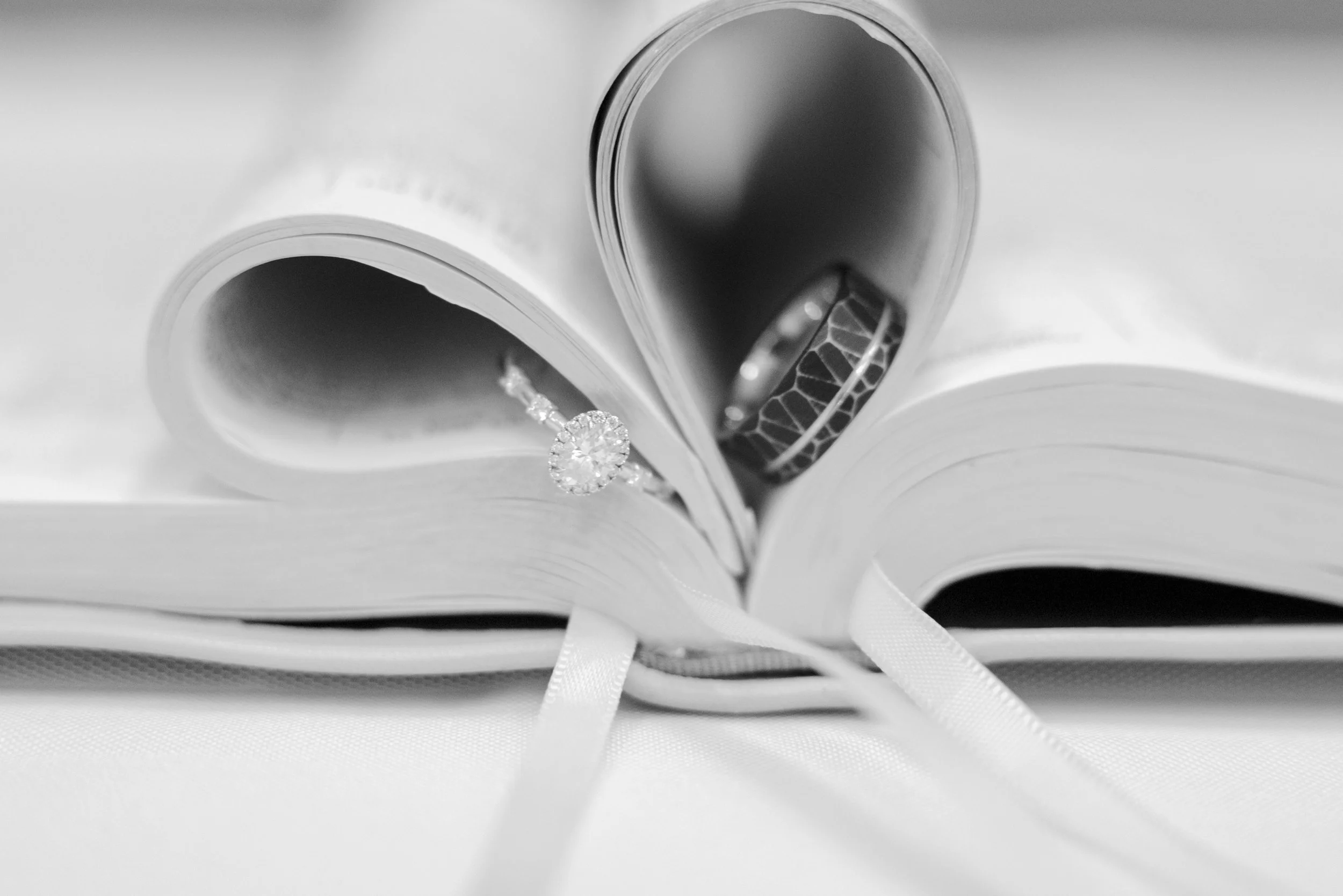 Open book with a diamond engagement ring and a patterned wedding band resting on its pages, in black and white.