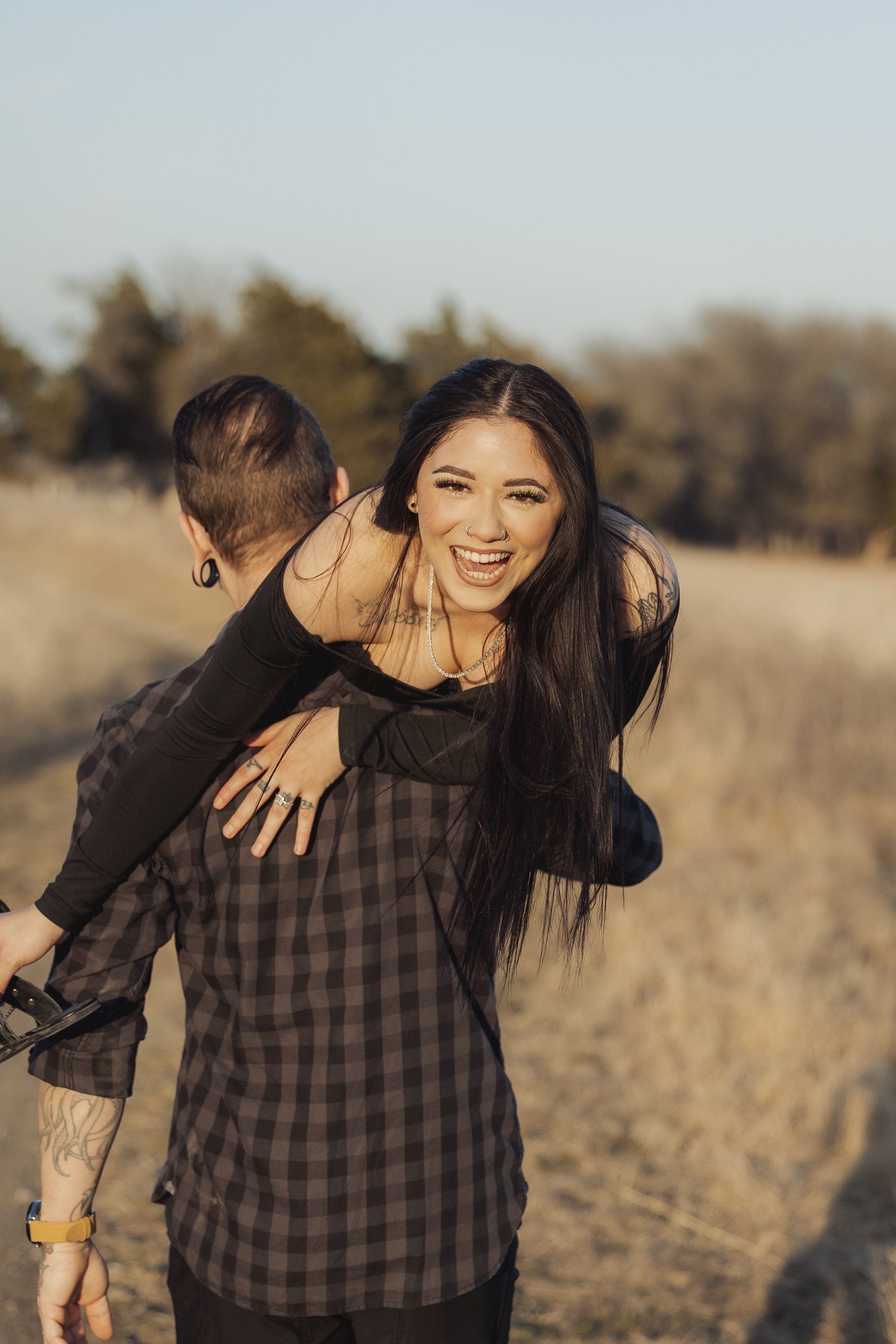 A woman with long black hair and tattoos being carried on a man's back outside in a field with trees in the background, smiling and looking at the camera.