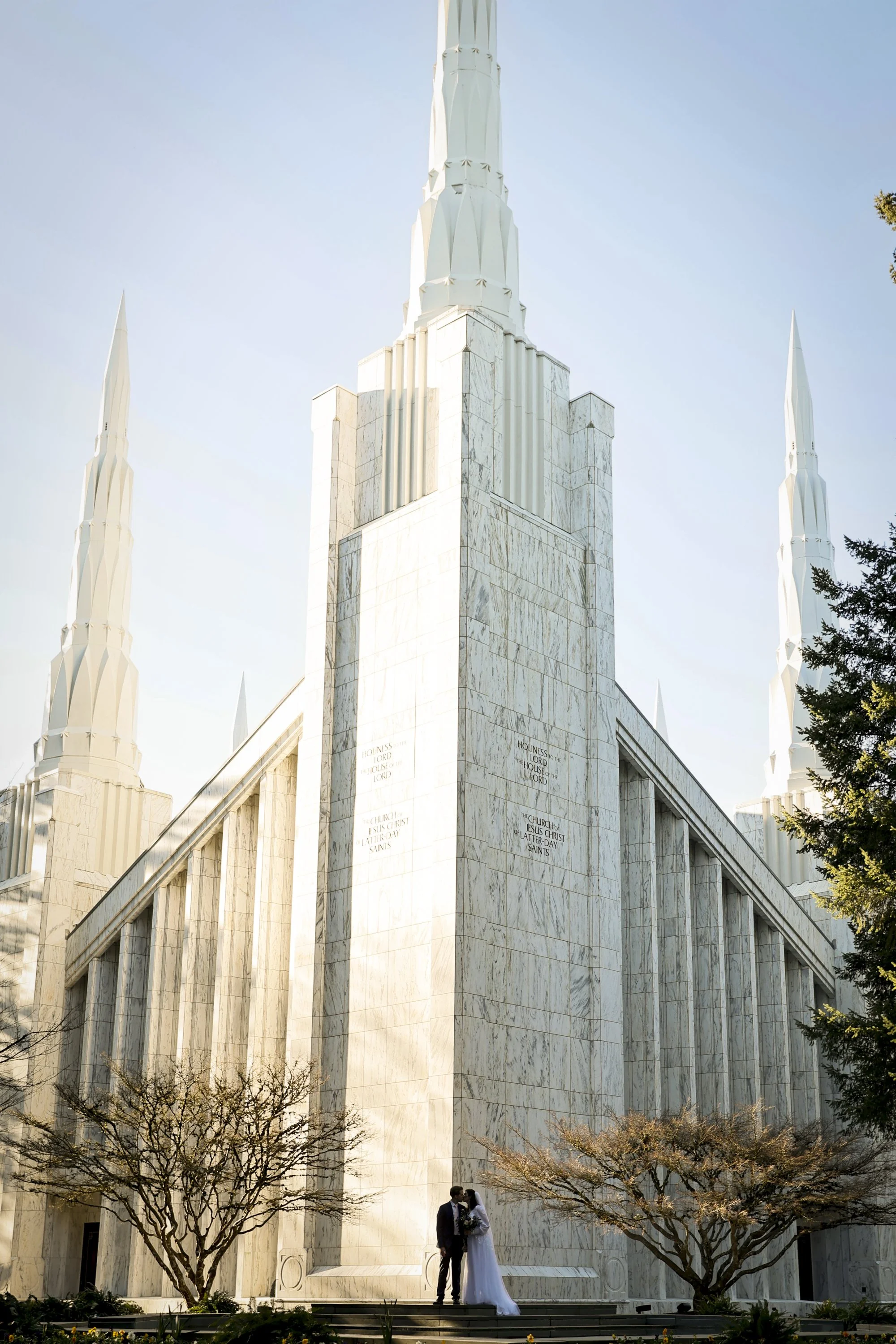 A bride and groom in wedding attire standing outside the Mormon (LDS) temple, which is made of white marble with tall spires, surrounded by leafless trees and partly cloudy sky.