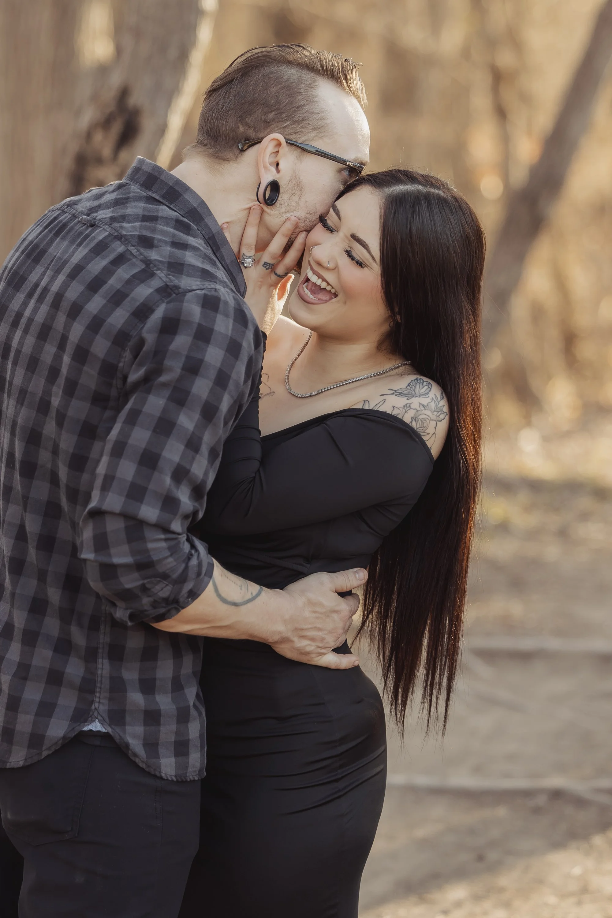 A couple sharing a kiss outdoors during sunset, with the man gently holding the woman, who is smiling with tattoos visible on her shoulder and wearing jewelry.