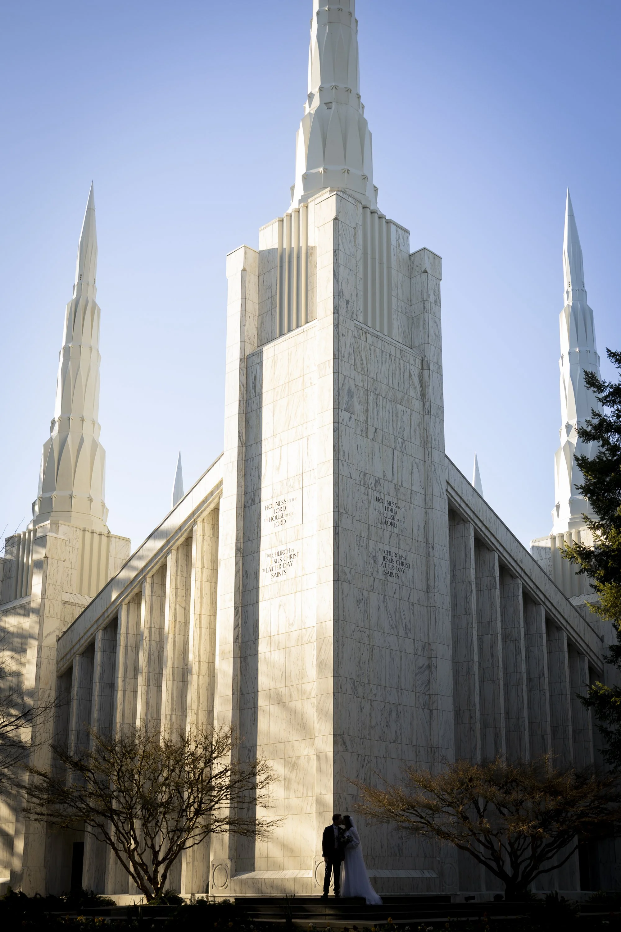 A bride and groom standing together outside the Portland Temple, a large white marble religious building with tall spires, during daytime.