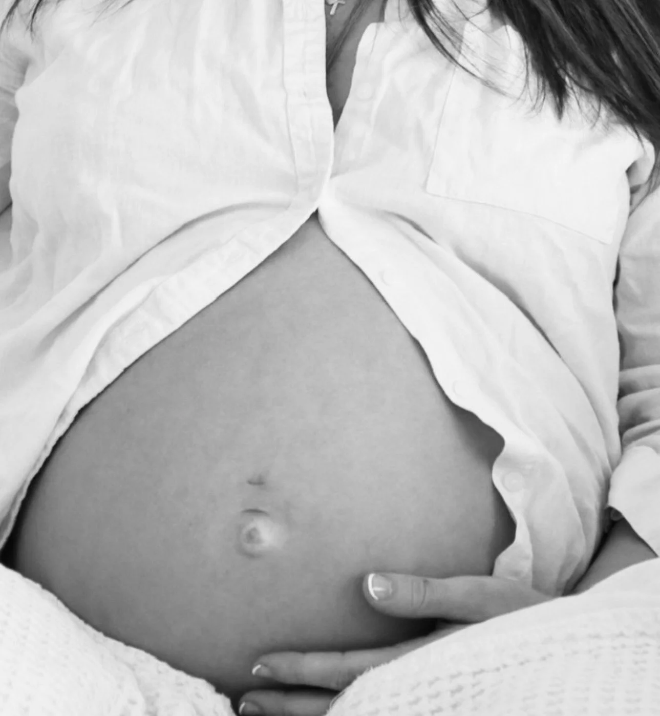 A black and white photo of a woman with her shirt unbuttoned, exposing her pregnant belly and navel, with one hand resting on her abdomen.