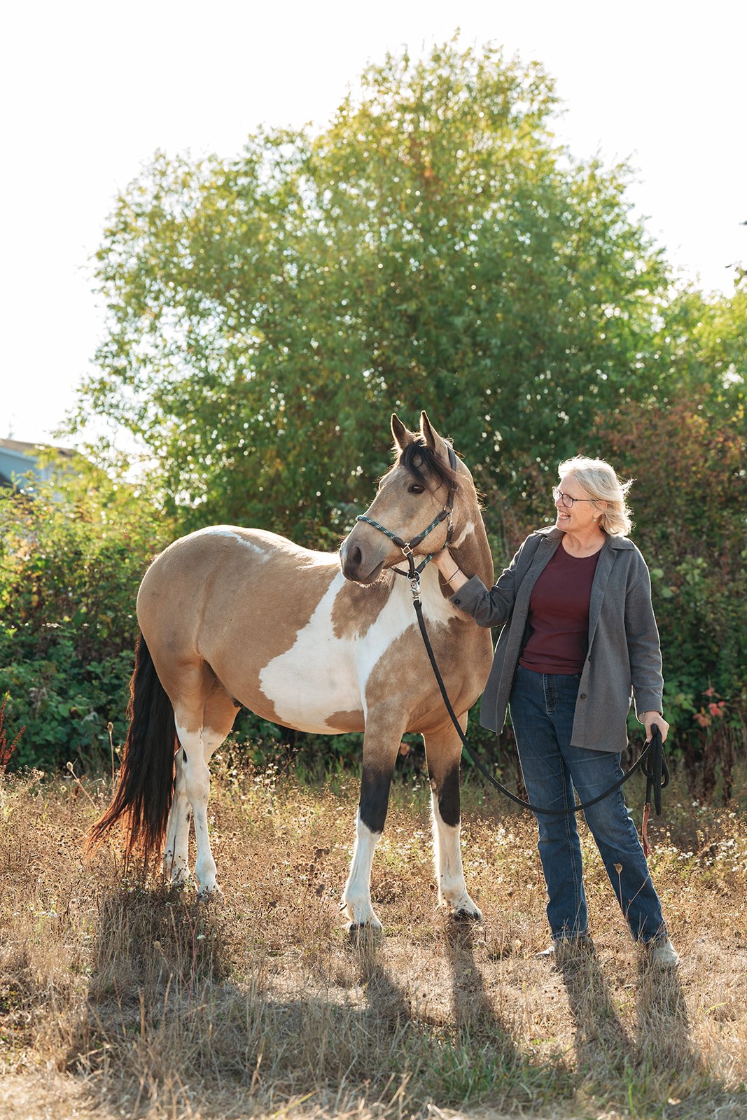 A woman with dressed in casual clothing standing outside with a  horse on an autumn day. The scene is set in a grassy area with trees in the background and sunlight filtering through the leaves.