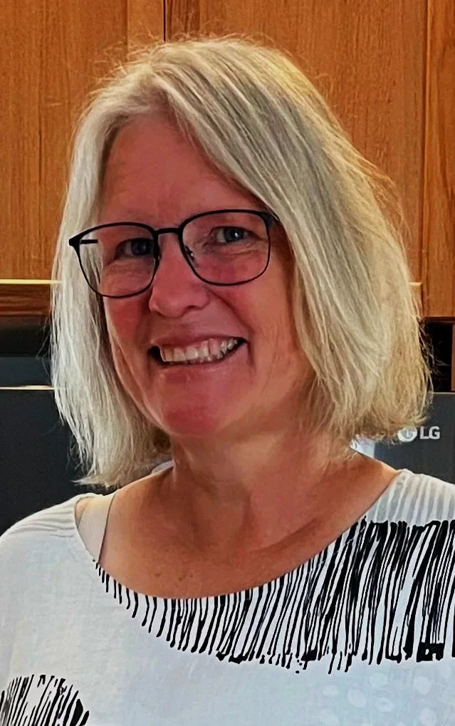 A woman with shoulder-length blonde hair, glasses, and a striped top smiling in an indoor setting with wooden paneling in the background.