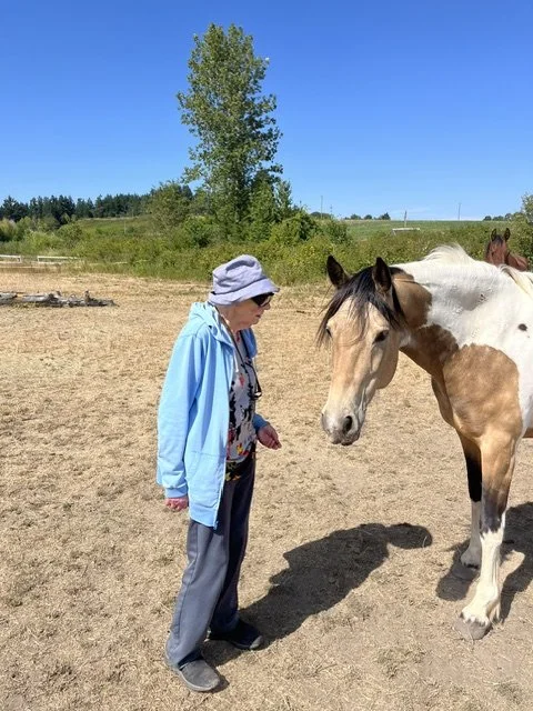 An elderly woman in a blue jacket and wide-brimmed hat standing on a dirt field, looking at a light-colored horse with brown patches.