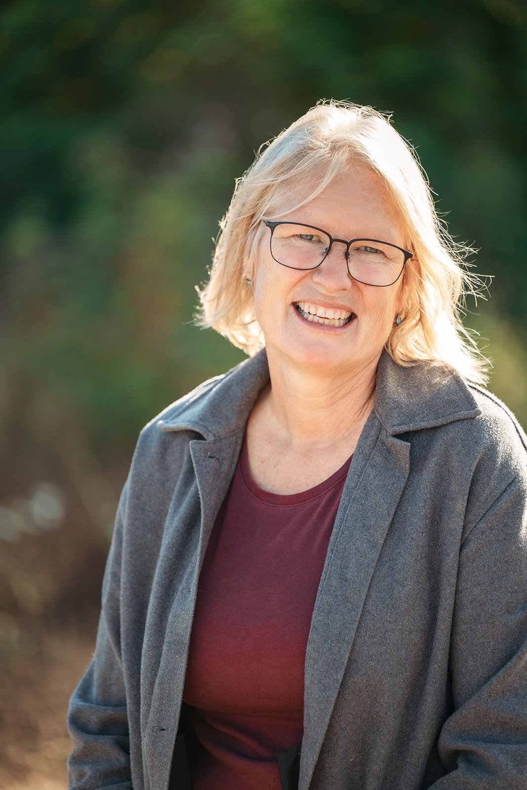 A smiling woman with glasses and short blonde hair, wearing a maroon shirt and a gray jacket, outdoors with blurred green foliage in the background.