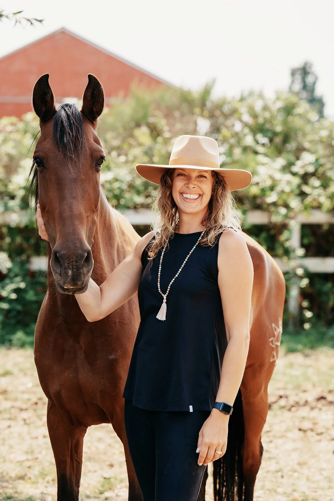 A woman smiling and wearing a large sunhat, black sleeveless top, and a beaded necklace, standing beside a brown horse outdoors with greenery and a building in the background.