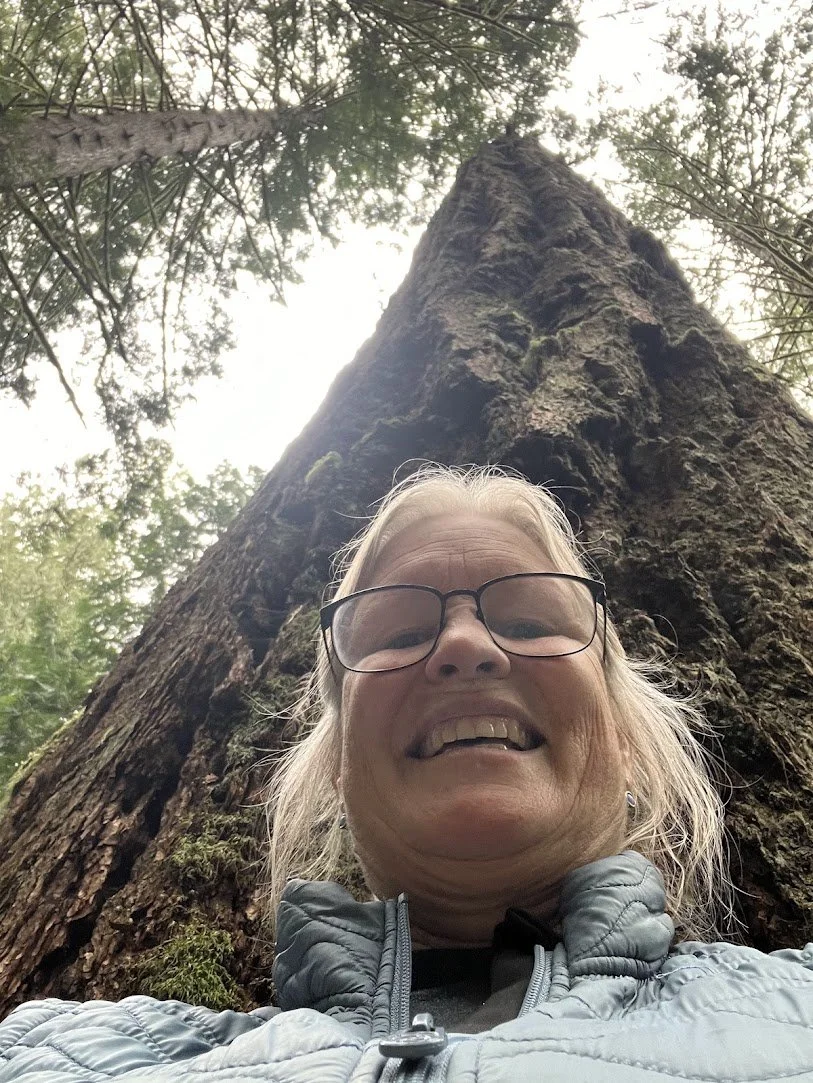 A woman with glasses smiling in a forest, standing in front of a tall tree trunk.