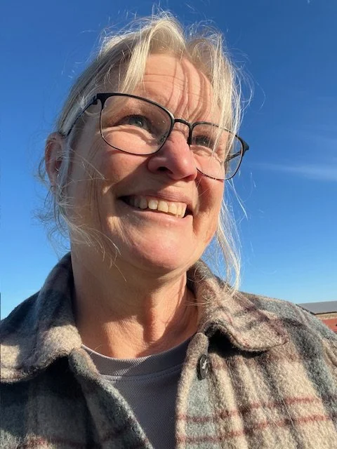 A smiling woman with gray hair and glasses outdoors on a sunny day, wearing a plaid jacket.