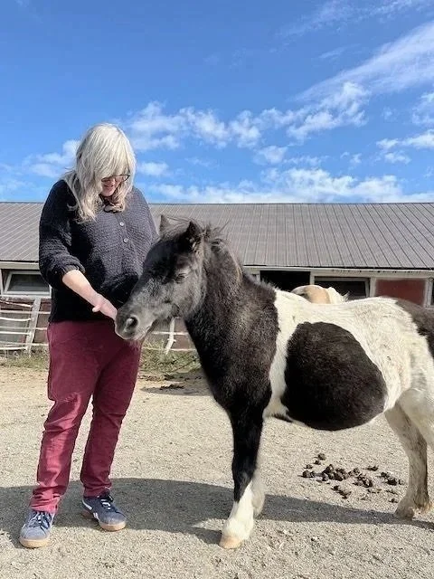 A woman with glasses and long  hair stands on a farm, gently touching a small black and white horse. The sky is partly cloudy with blue skies, and there is a barn and fencing in the background.