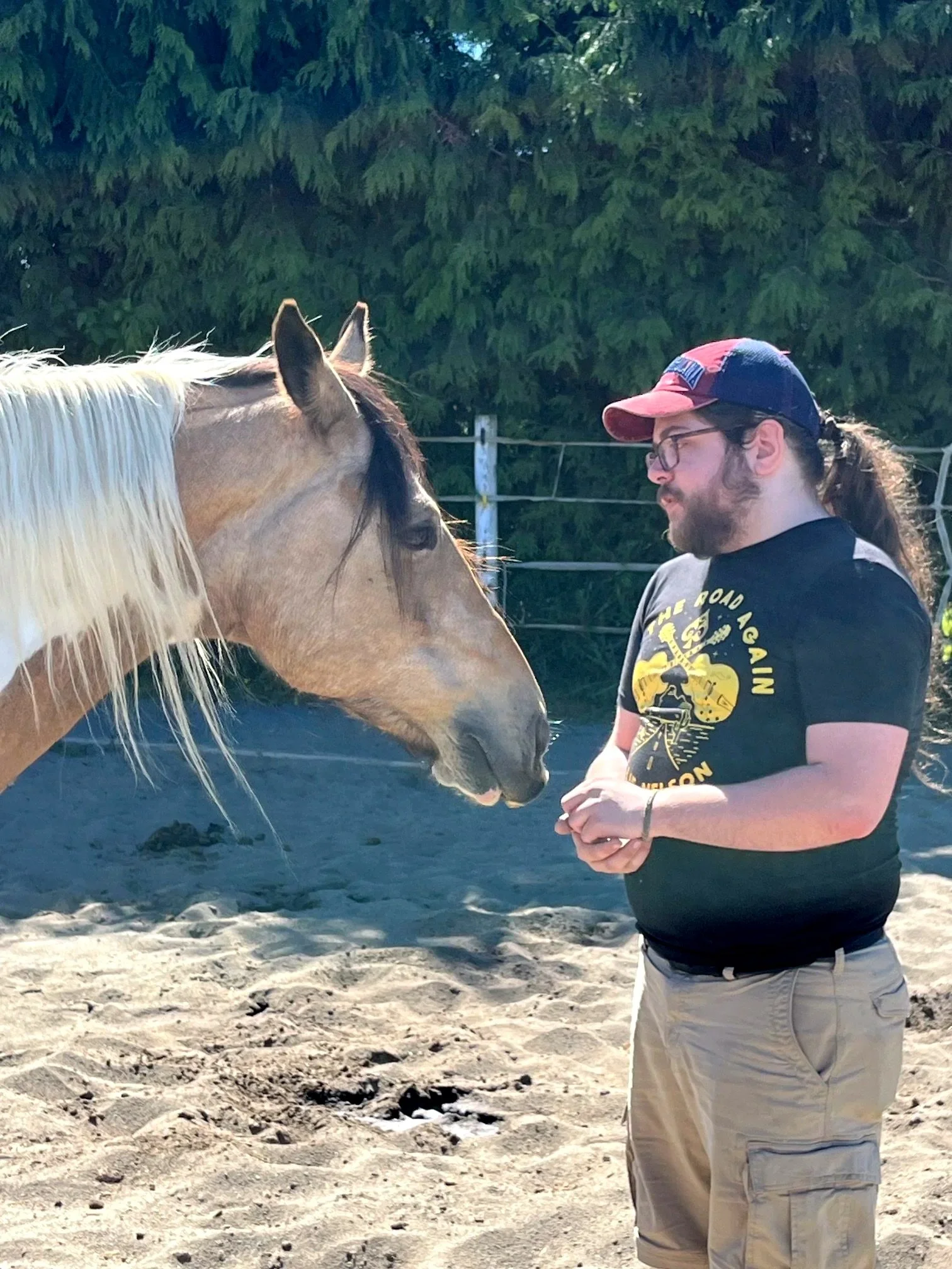 A man with glasses, a beard, and a ponytail, wearing a baseball cap and a black T-shirt facing a light-colored horse with dark mane, in an outdoor area with trees in the background.