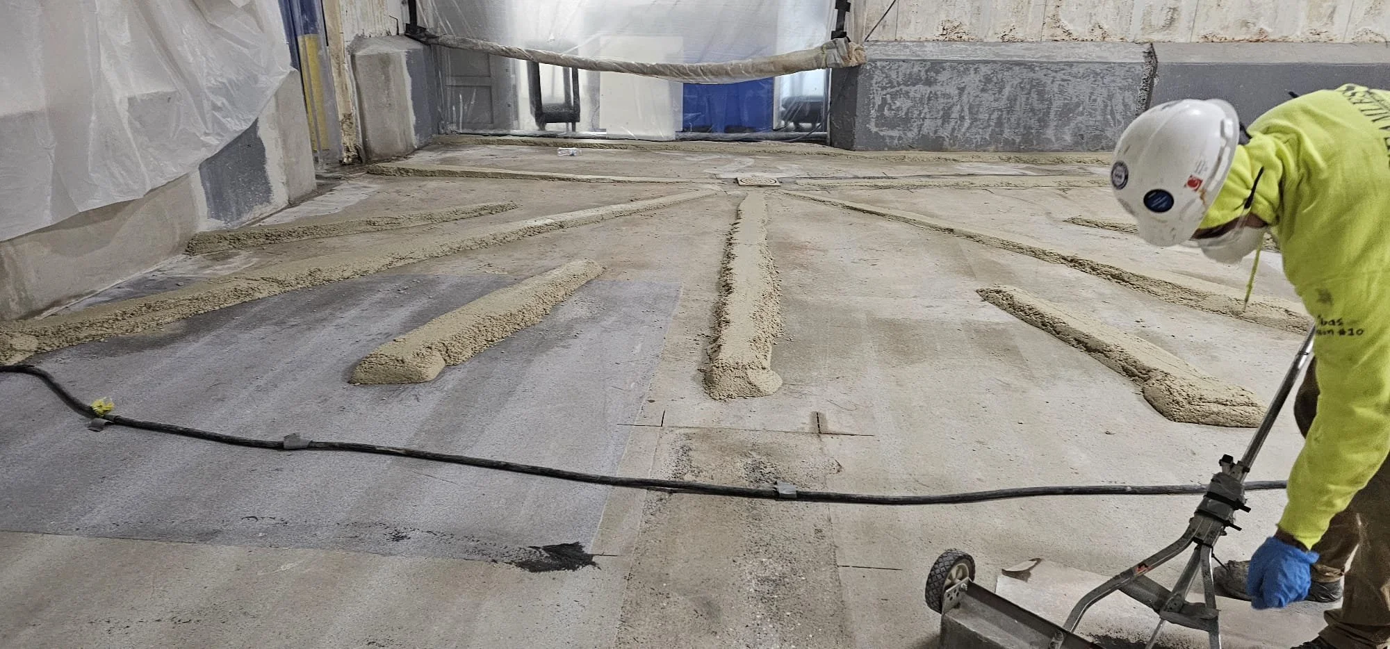A construction worker with a white helmet and yellow safety vest is installing or repairing concrete flooring, with lines of fresh concrete being smoothed out.