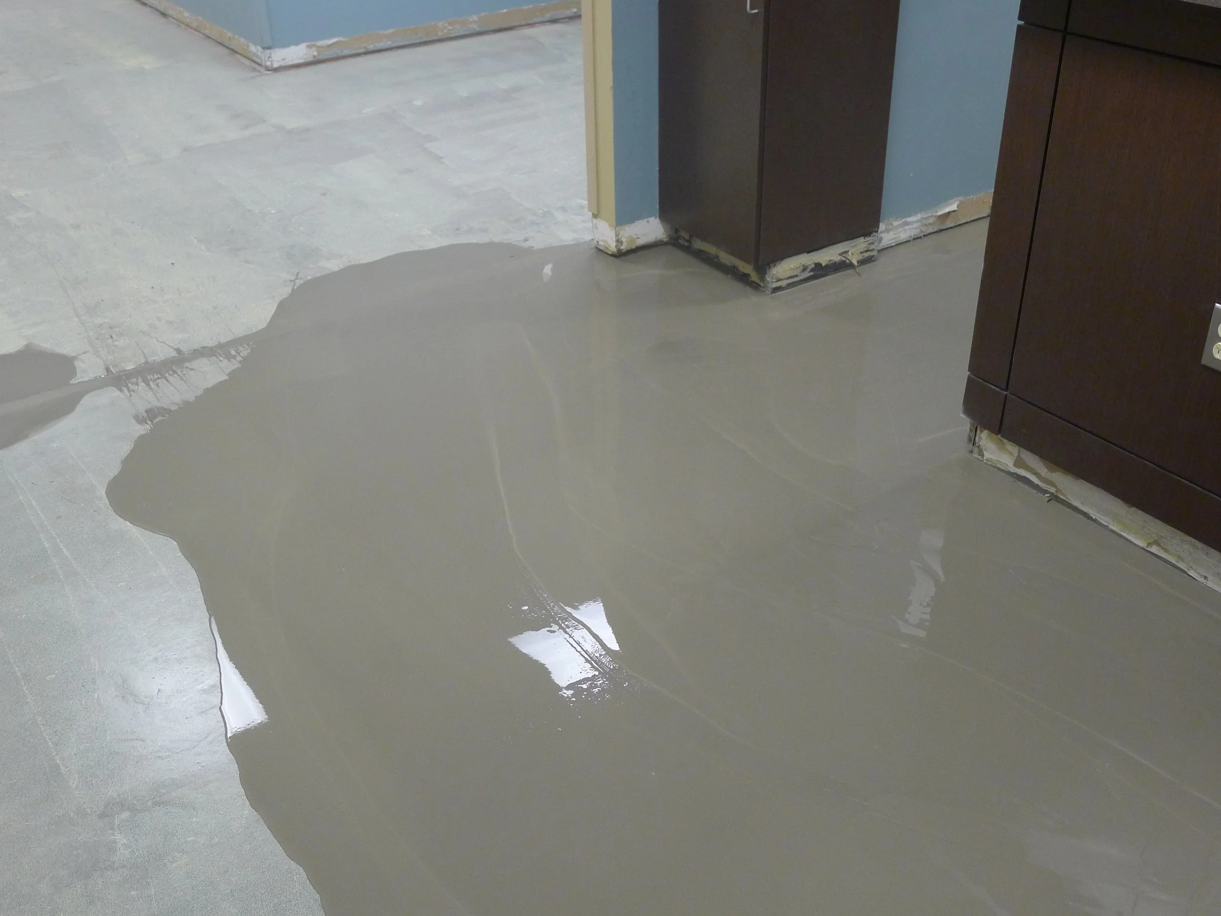 Wet concrete spilled on light gray tile floor near dark brown cabinets in a room under renovation.
