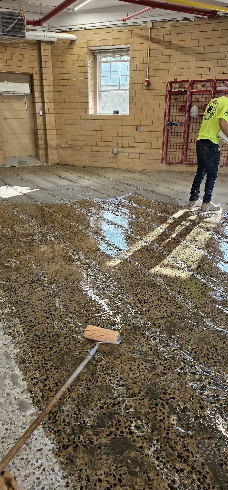 A person in a yellow shirt working on a concrete floor with a roller, in a room with brick walls and a window. The floor is in the process of being sealed or coated.