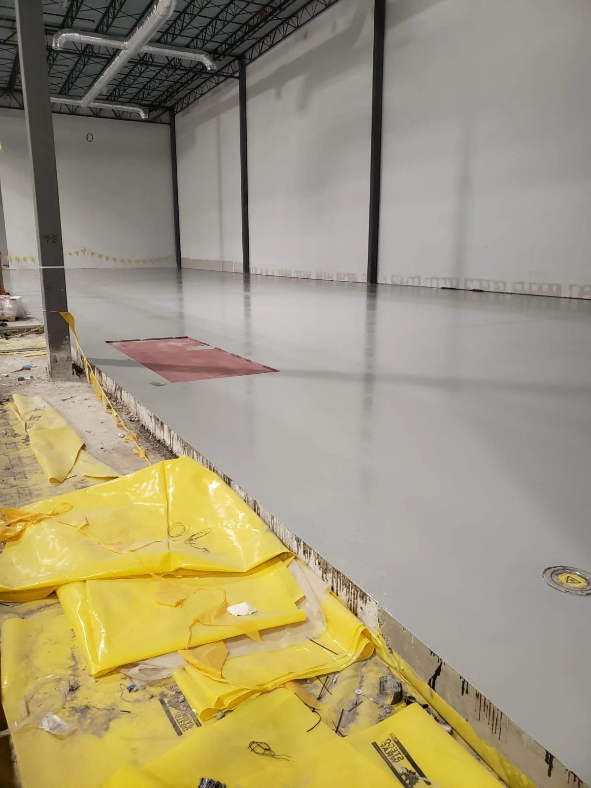 Indoor construction site with a freshly poured concrete floor, yellow caution tarp on the floor, and red access hatch in the concrete.