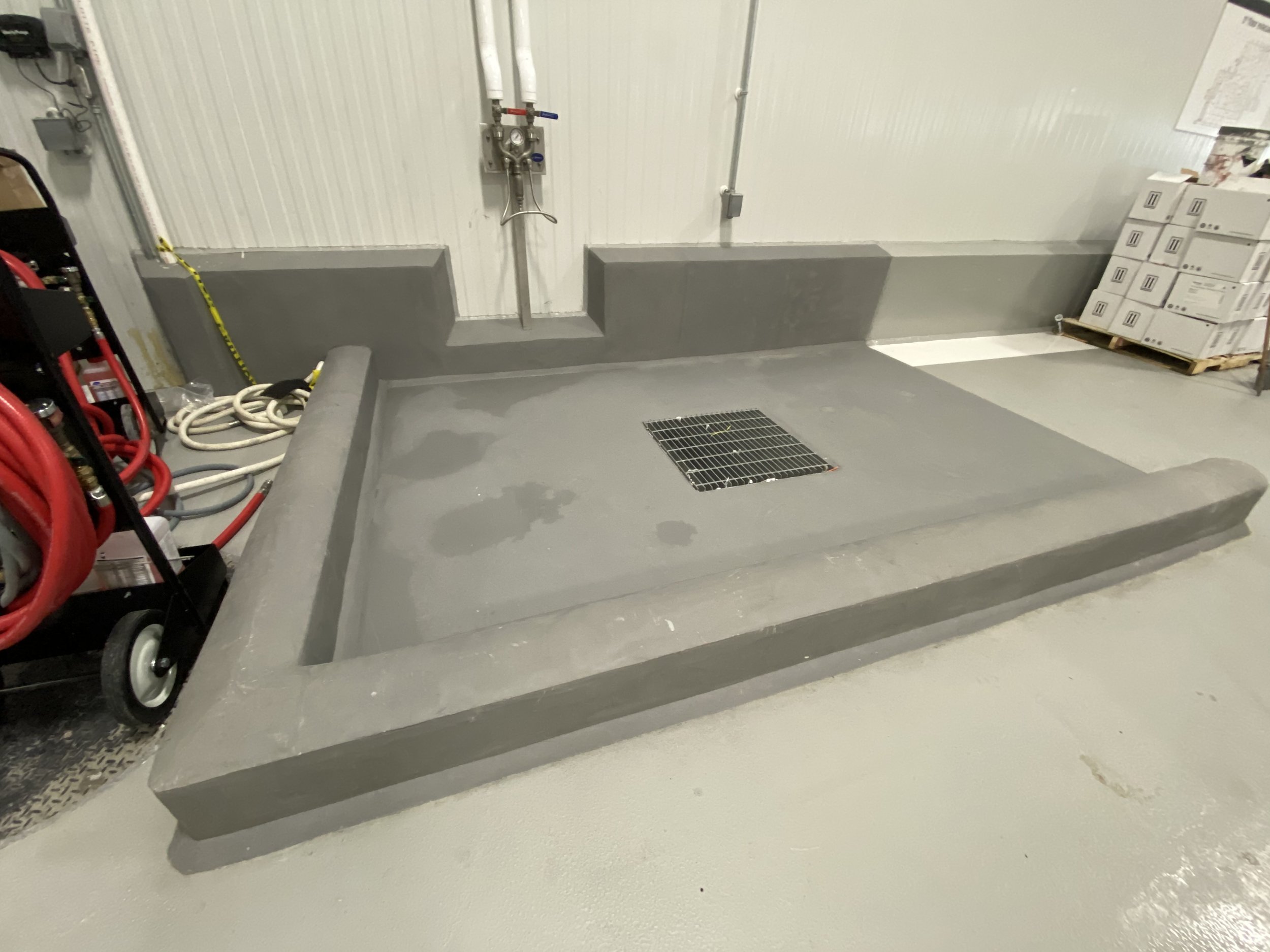 Concrete wash basin with a drain and gray walls, located in an industrial or utility area, with cleaning supplies, hoses, and storage boxes nearby.