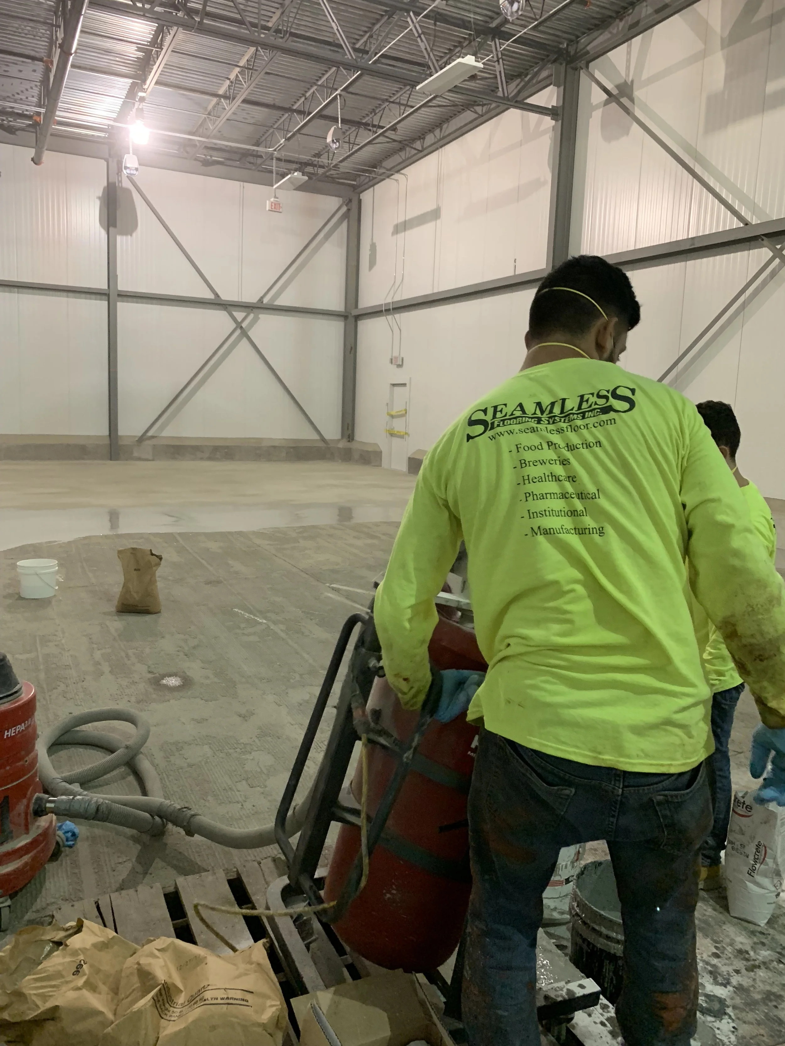 Construction workers in a large warehouse space with unfinished floors and high metal ceiling, working on a project.