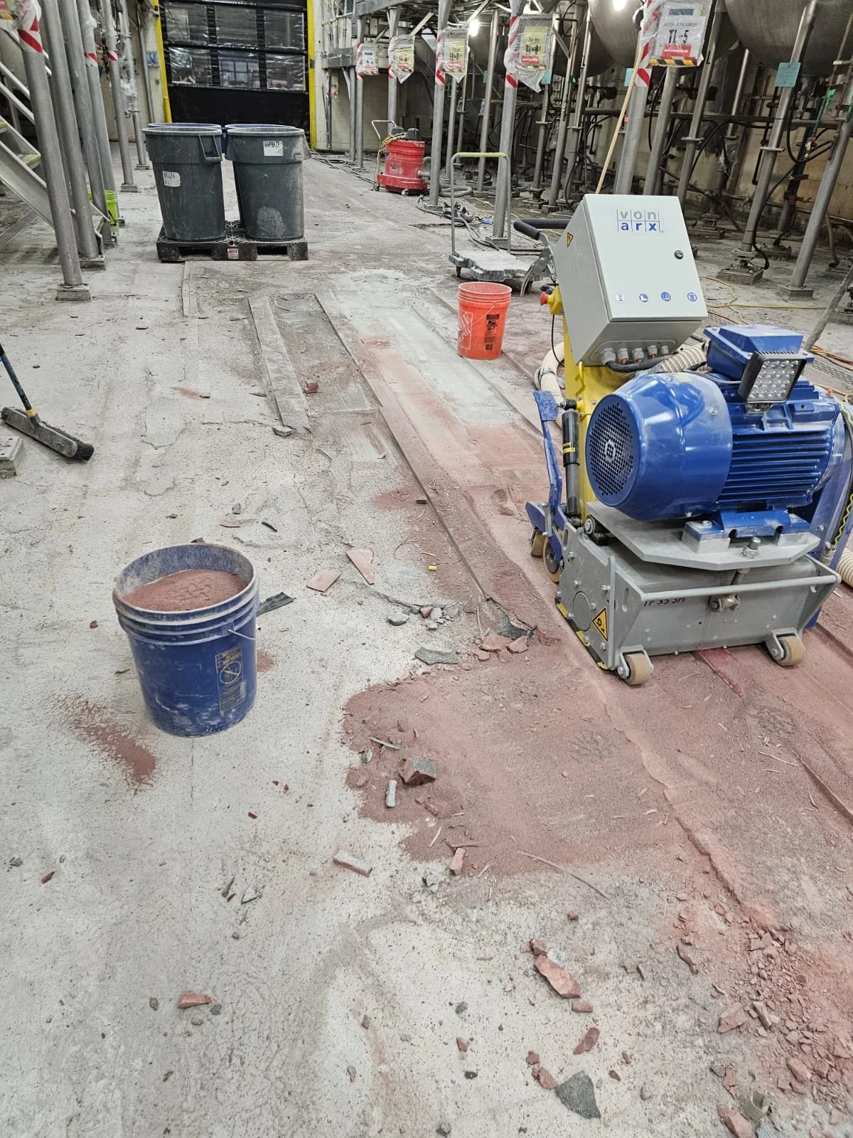 Interior construction site with dust, debris, construction tools, and equipment, including a large blue machine, orange buckets, and metal scaffolding.