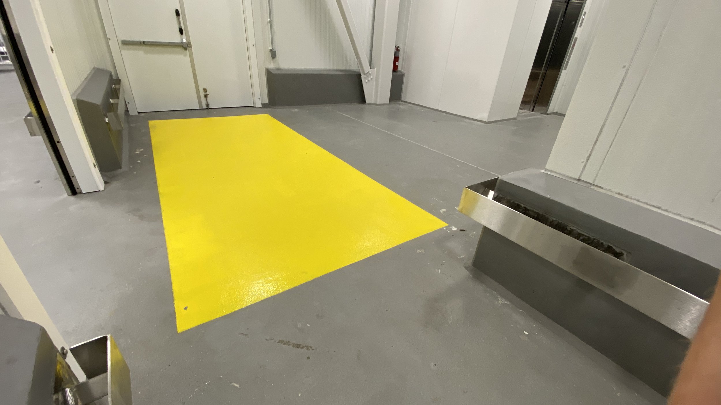 The image shows a concrete floor with a bright yellow rectangular strip painted on it. To the left, there is a waist-height metal trash can, and to the right, there is a metallic water fountain or trough. In the background, there are doors and part o