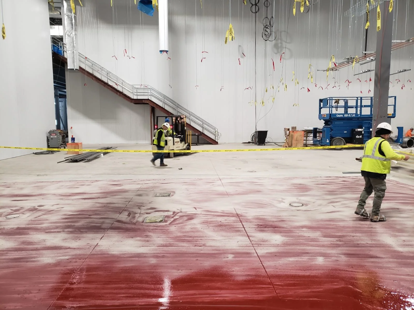 Construction workers in yellow safety vests and helmets working inside a large industrial building with a red floor, yellow caution tape, and construction equipment.