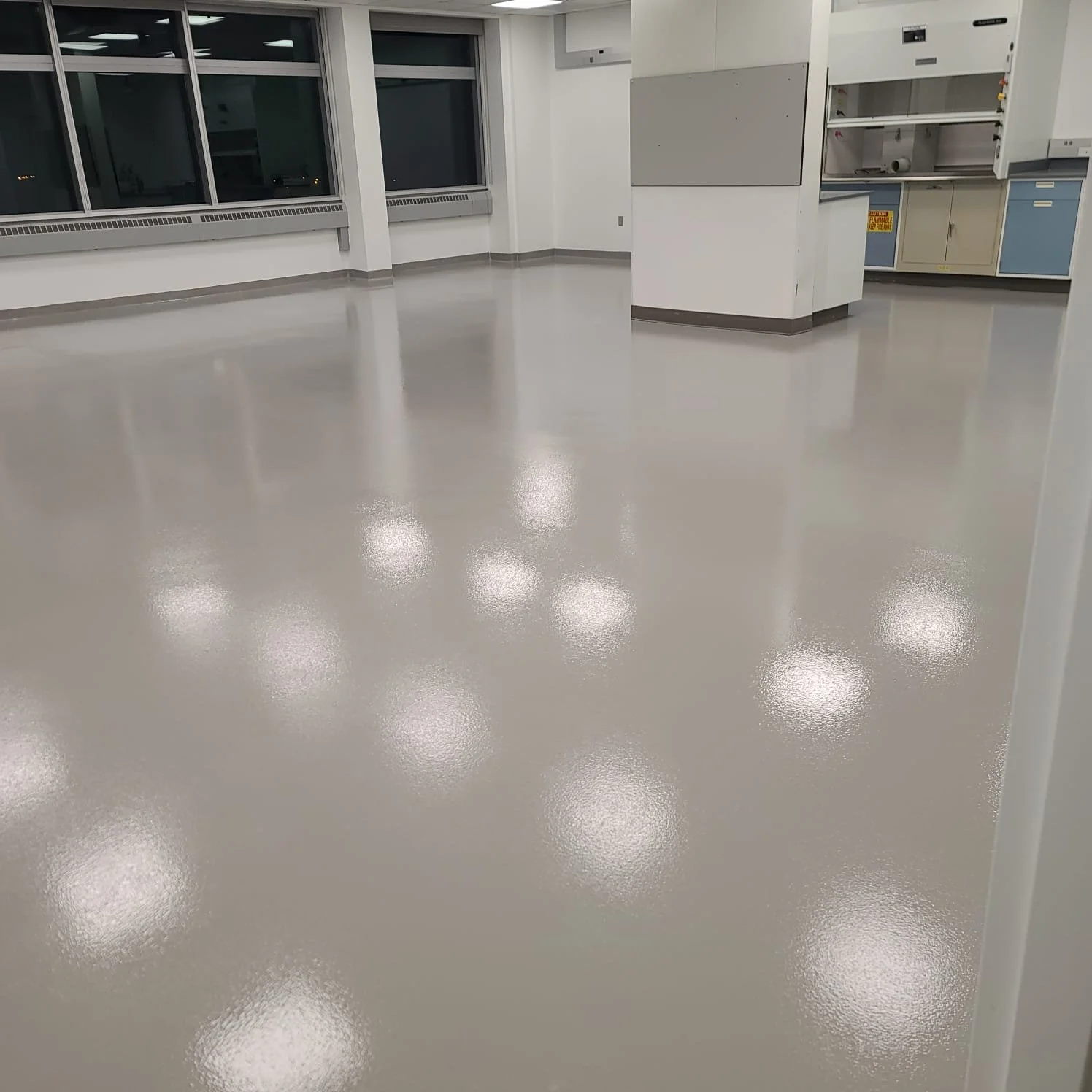 Empty laboratory room with large windows, bright overhead lights, and white walls, featuring blue and beige cabinets and countertops.