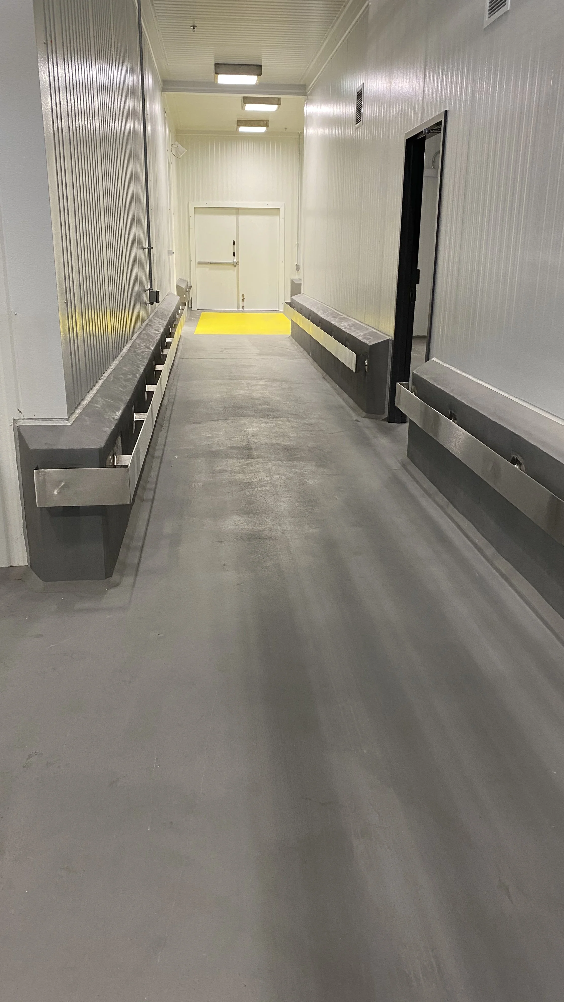 Empty industrial or loading dock hallway with gray concrete floor, white metal walls, stainless steel bumper guards, and a yellow safety strip at the end near double doors.