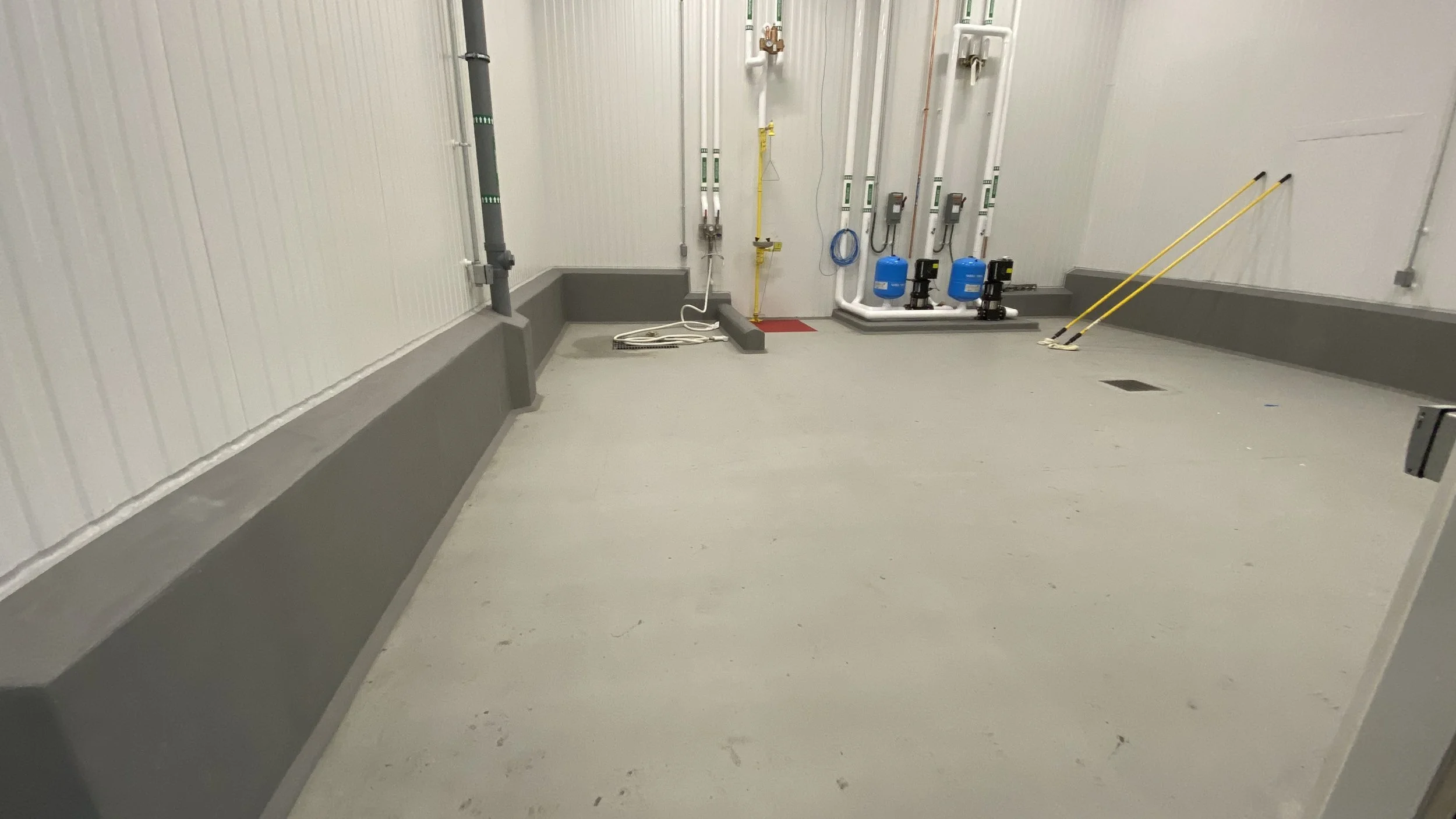 An industrial utility or mechanical room with pipes, meters, and blue tanks installed on the wall; yellow poles leaning against the wall; and a floor drain.