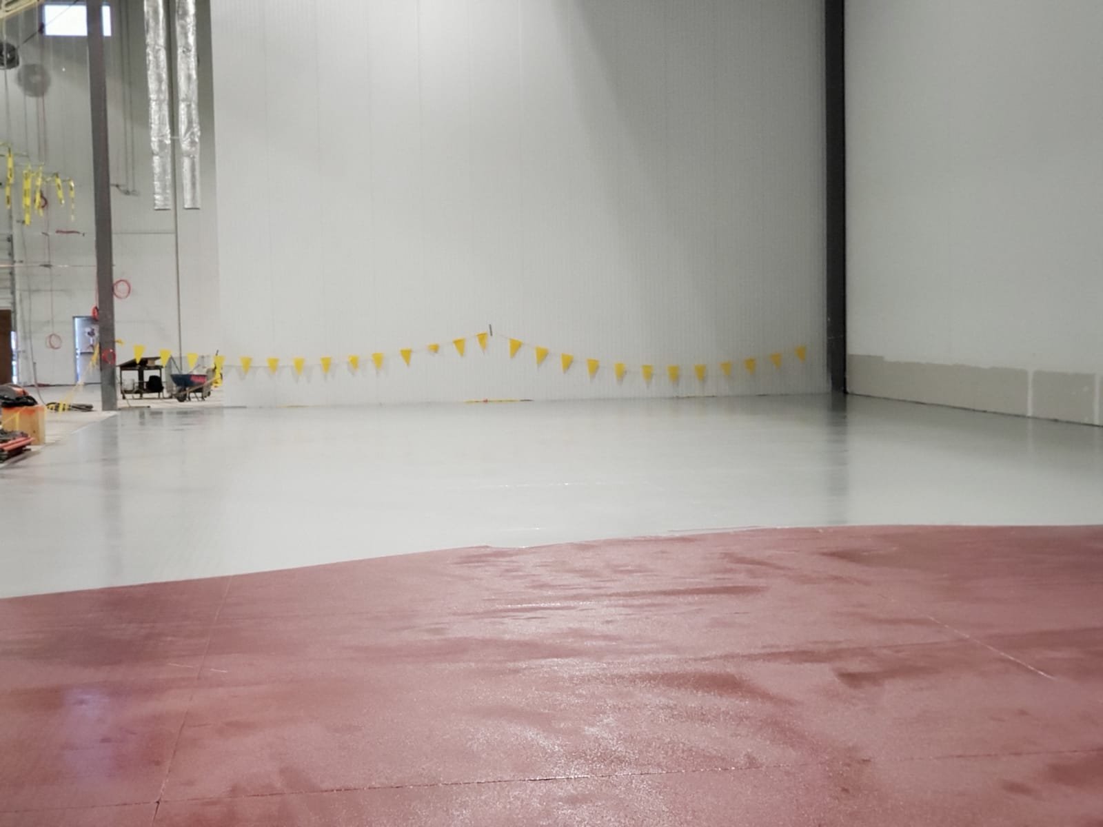 Interior view of an industrial or warehouse space with a red painted floor in foreground and a large white wall with yellow caution tape in the background.