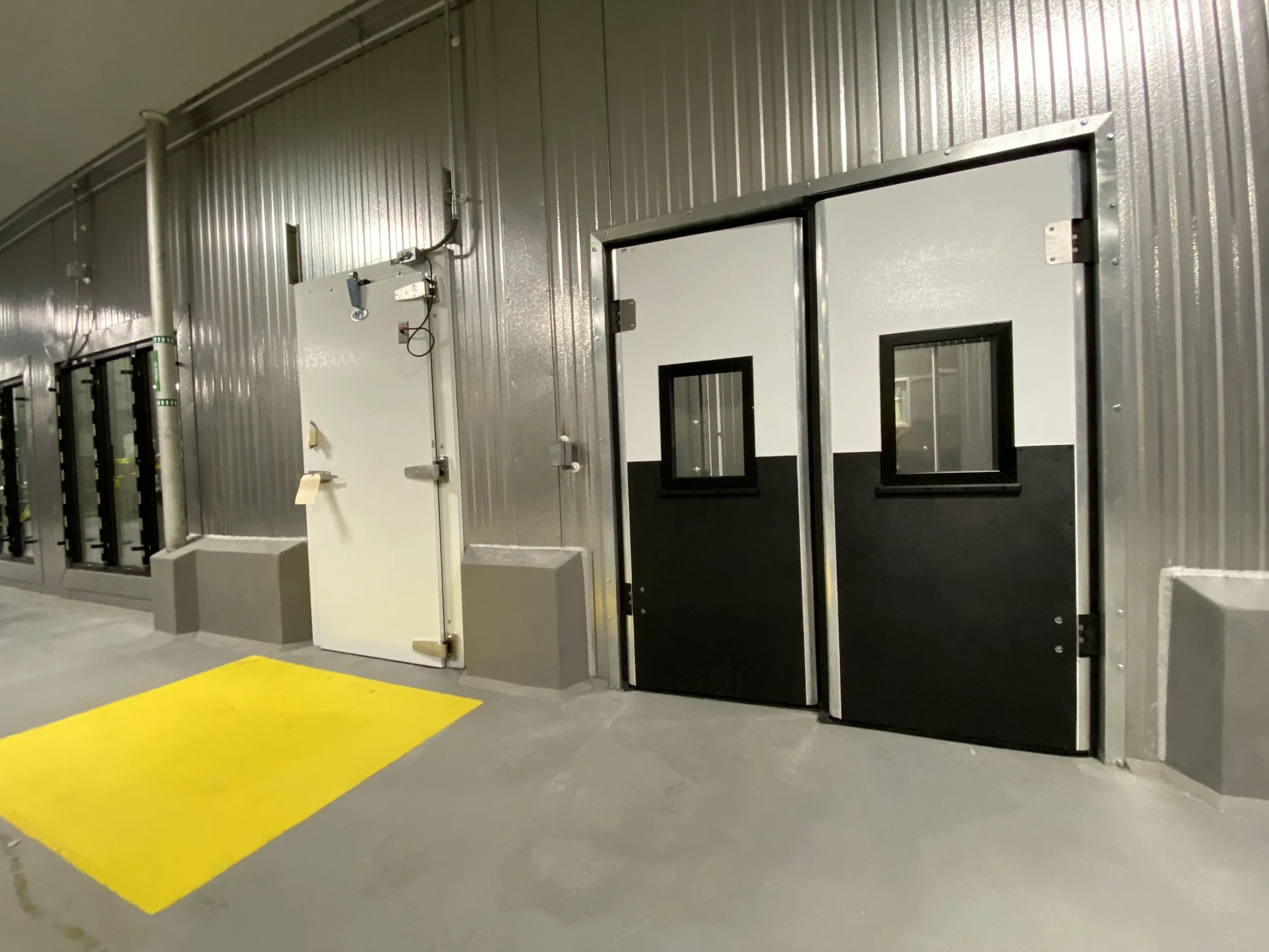 Industrial door with windows and yellow safety mat on concrete floor in a metal-walled room.