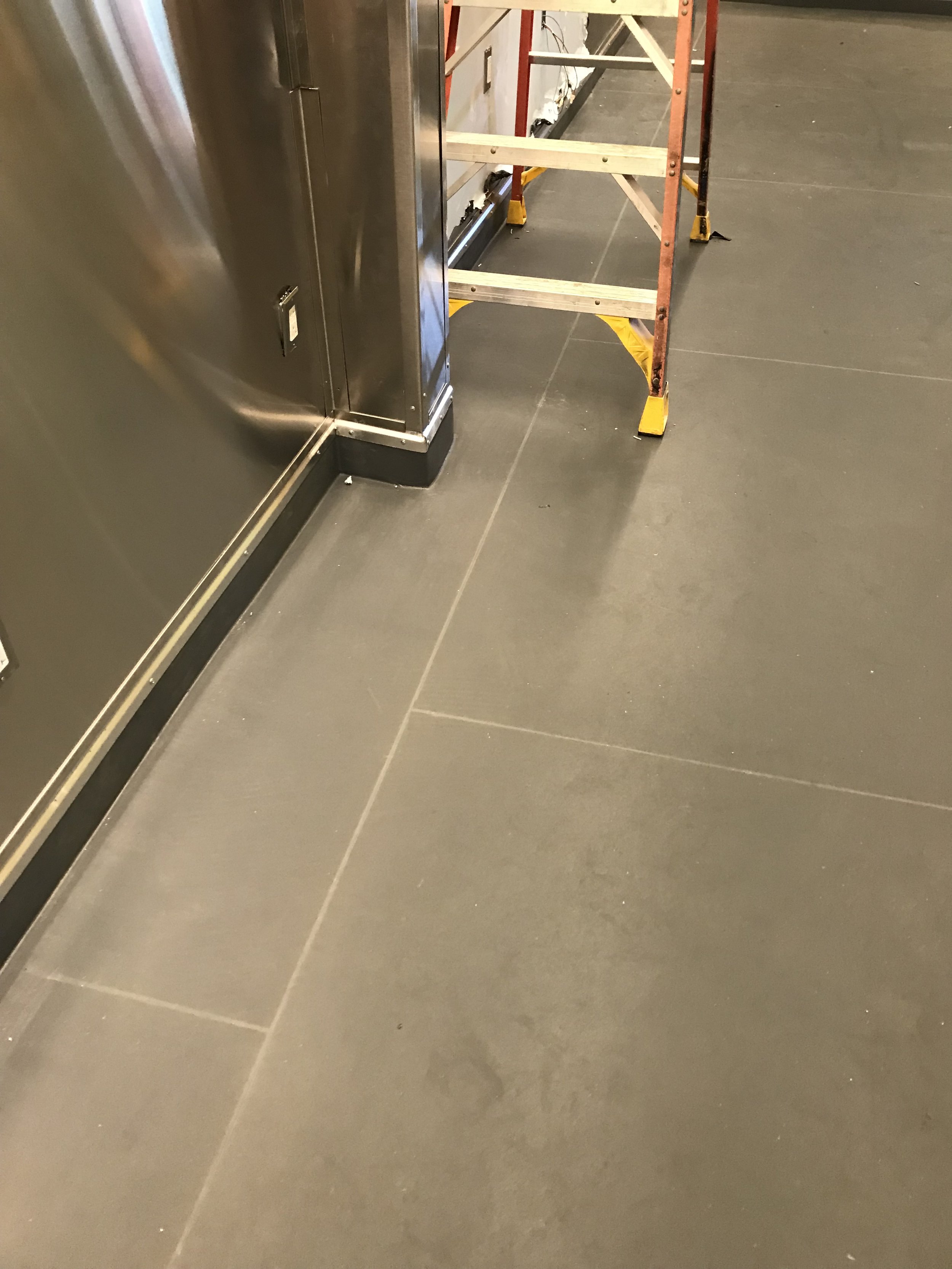 Part of a kitchen or industrial setting with a stainless steel appliance, a concrete or stone tile floor, and a wooden and metal step ladder.