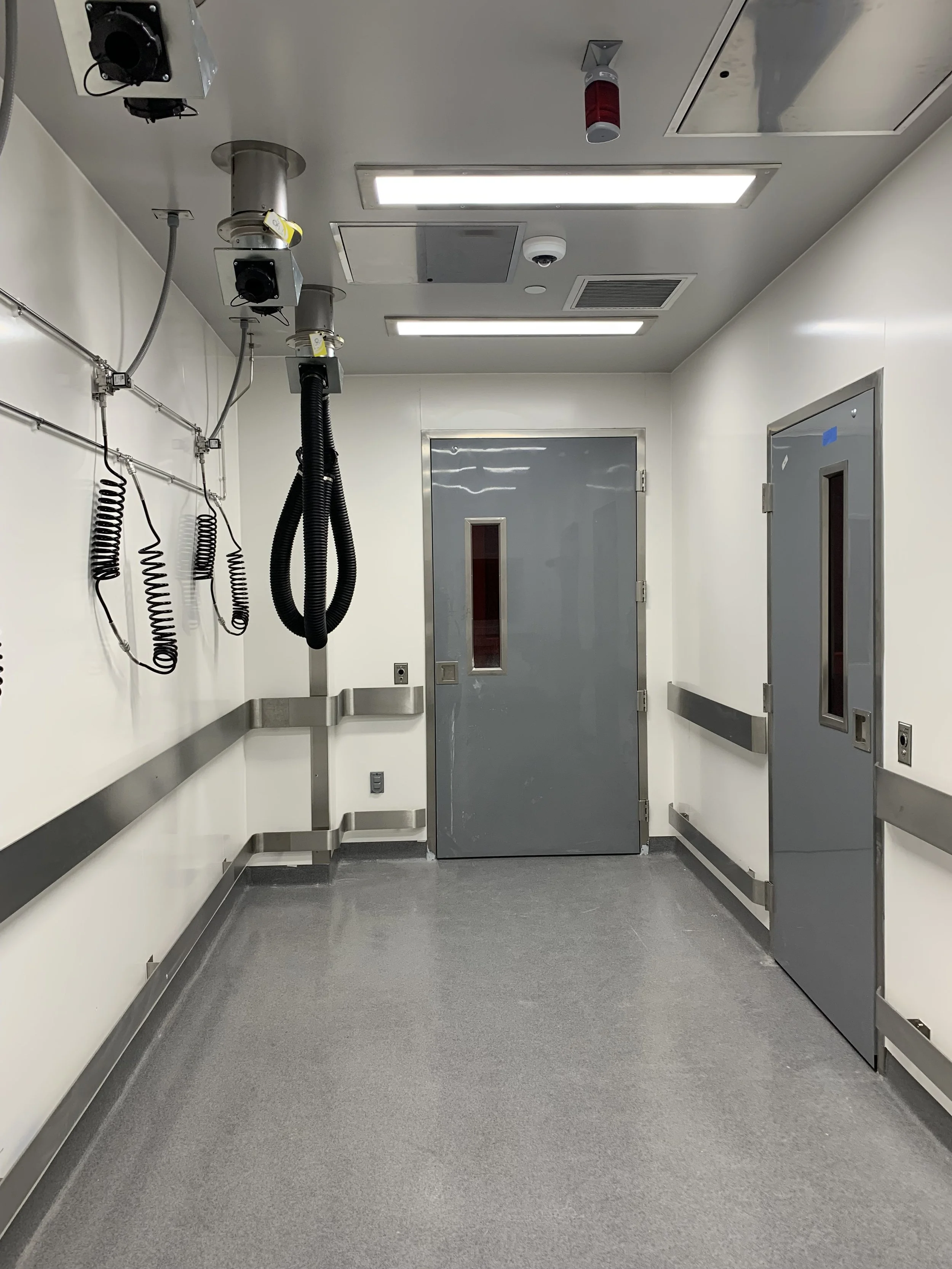 An empty hospital corridor or room with gray doors, metal handrails, ceiling-mounted medical equipment, and bright overhead lighting.