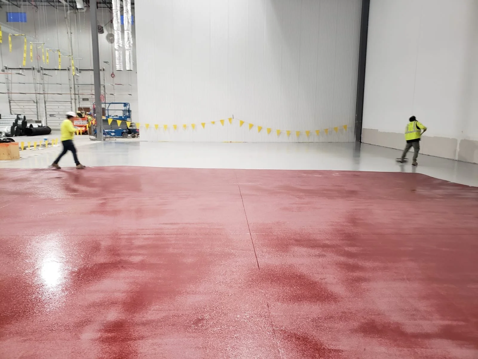 Interior of an industrial or warehouse space with a red epoxy floor and two workers in yellow safety vests. The space has high ceilings with construction equipment and materials visible in the background.